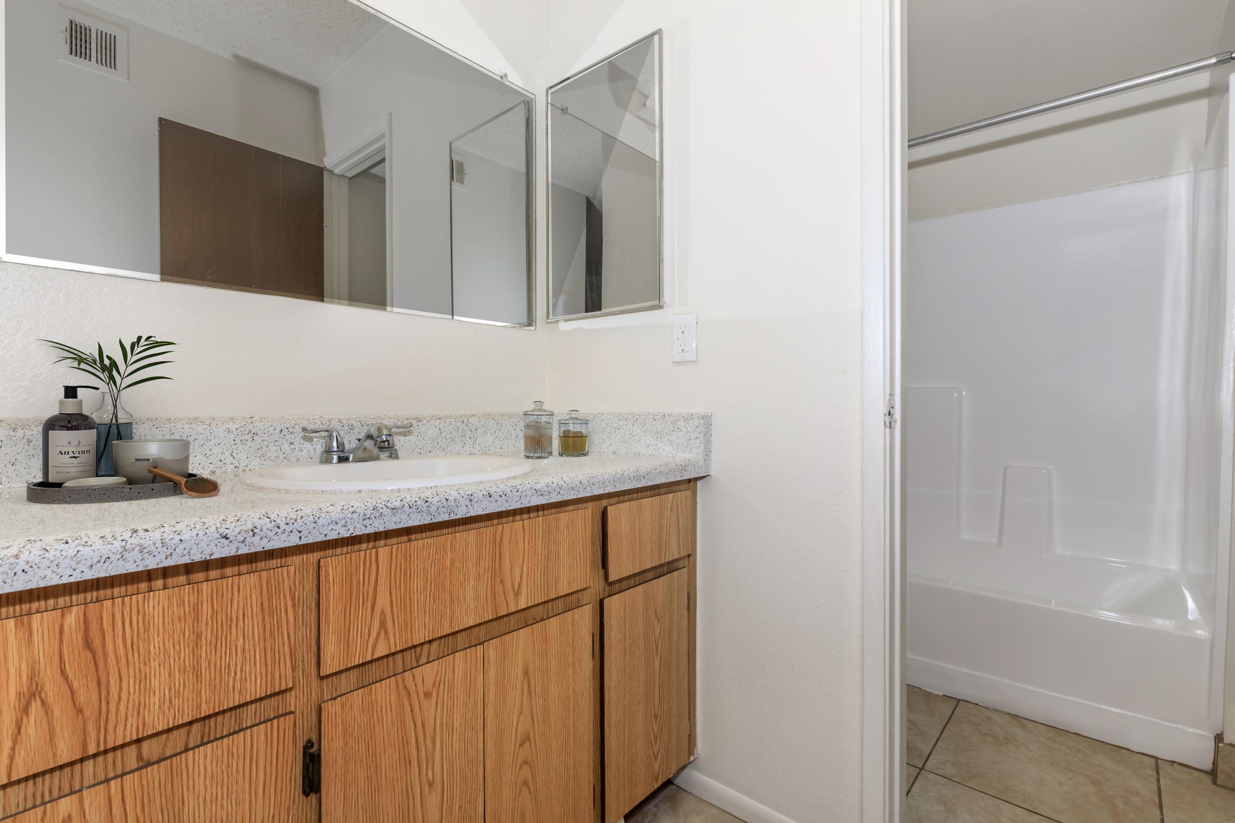 A bathroom featuring a light-colored countertop with two sinks, wooden cabinetry below, and three mirrors above. To the right, there's a bathtub with a shower curtain. Natural light enhances the clean, minimalist design, showcasing beige tiles and subtle decorative elements.