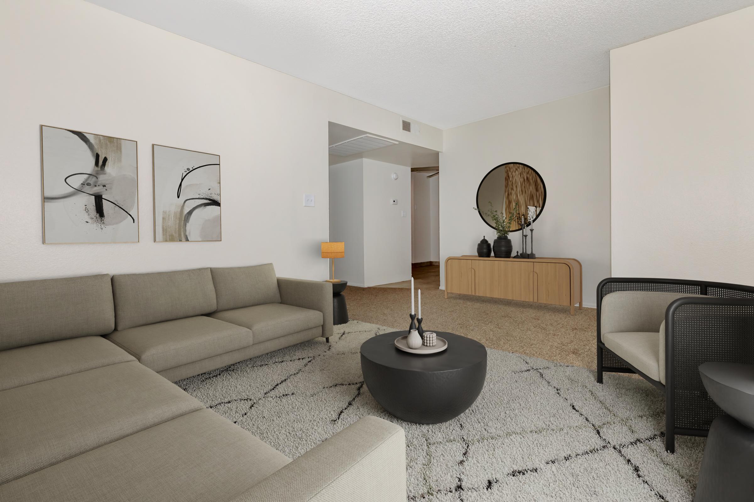 A modern living room featuring a light-colored sectional sofa, two contemporary black chairs, and a round coffee table. The space includes abstract wall art, a large circular mirror, and a side table with a lamp. The floor is covered with a plush, patterned rug, creating a cozy and stylish atmosphere.