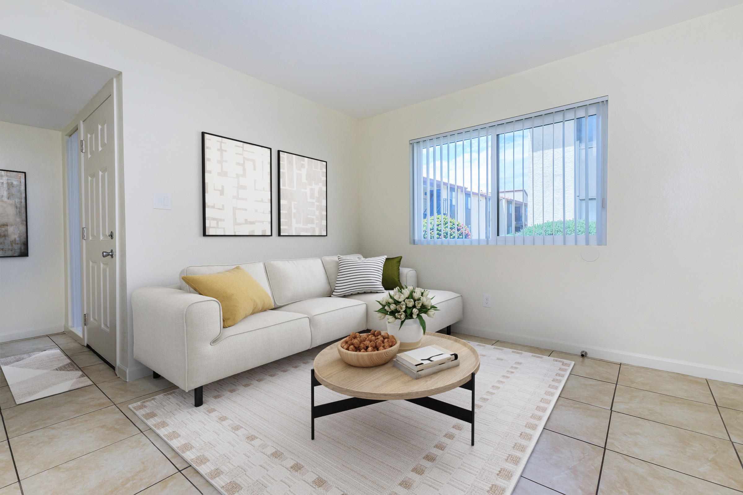 A cozy living room with a light beige sofa adorned with yellow and striped cushions. A round wooden coffee table sits in the center, holding a bowl of nuts and a small vase of flowers. Natural light streams in through a window with horizontal blinds, illuminating the soft cream walls and tiled floor.