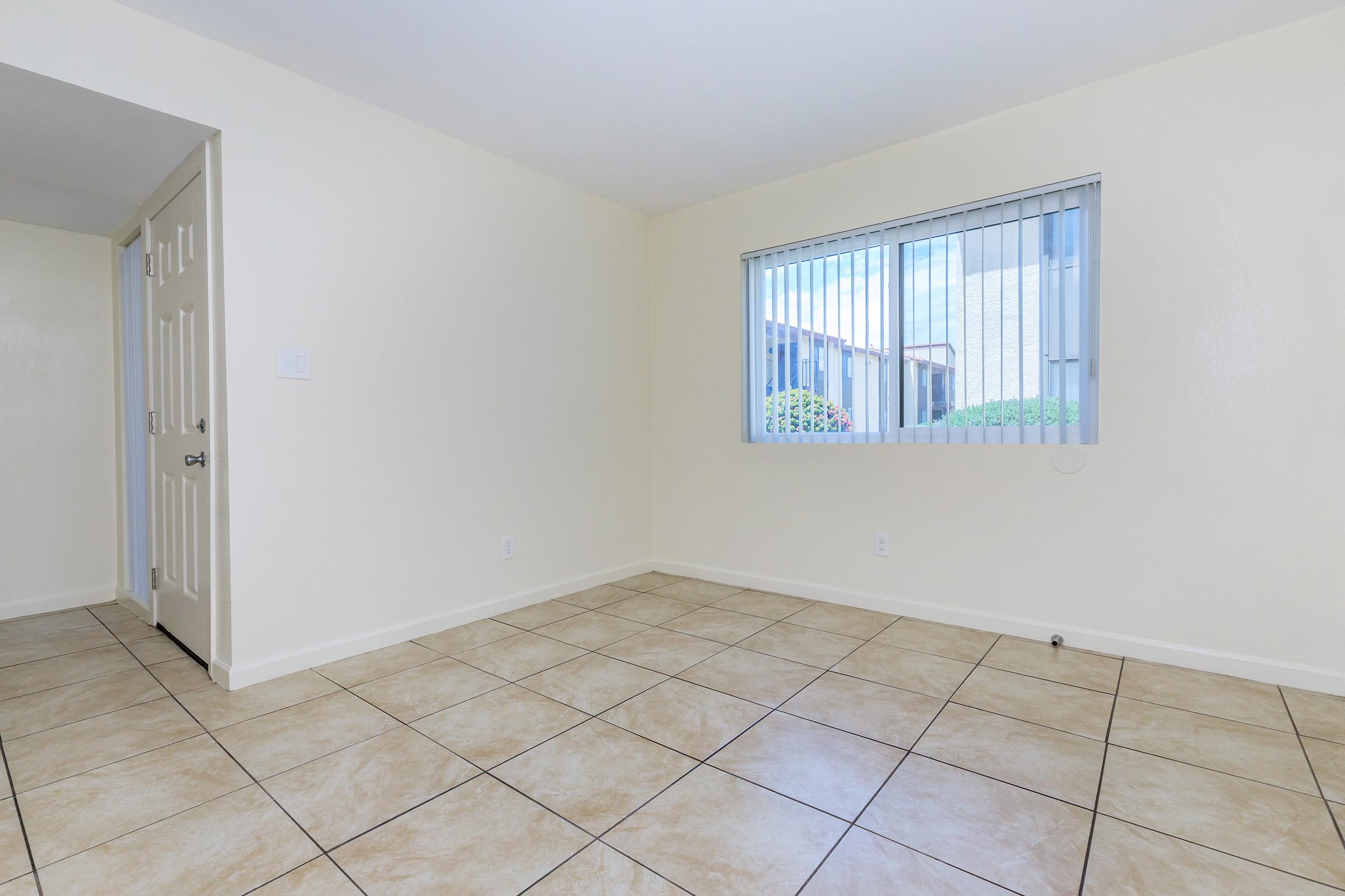 Empty room featuring light-colored walls and tiled flooring. A window with vertical blinds allows natural light, showcasing a view of greenery outside. The door on the left leads to a hallway, creating a sense of openness and space.