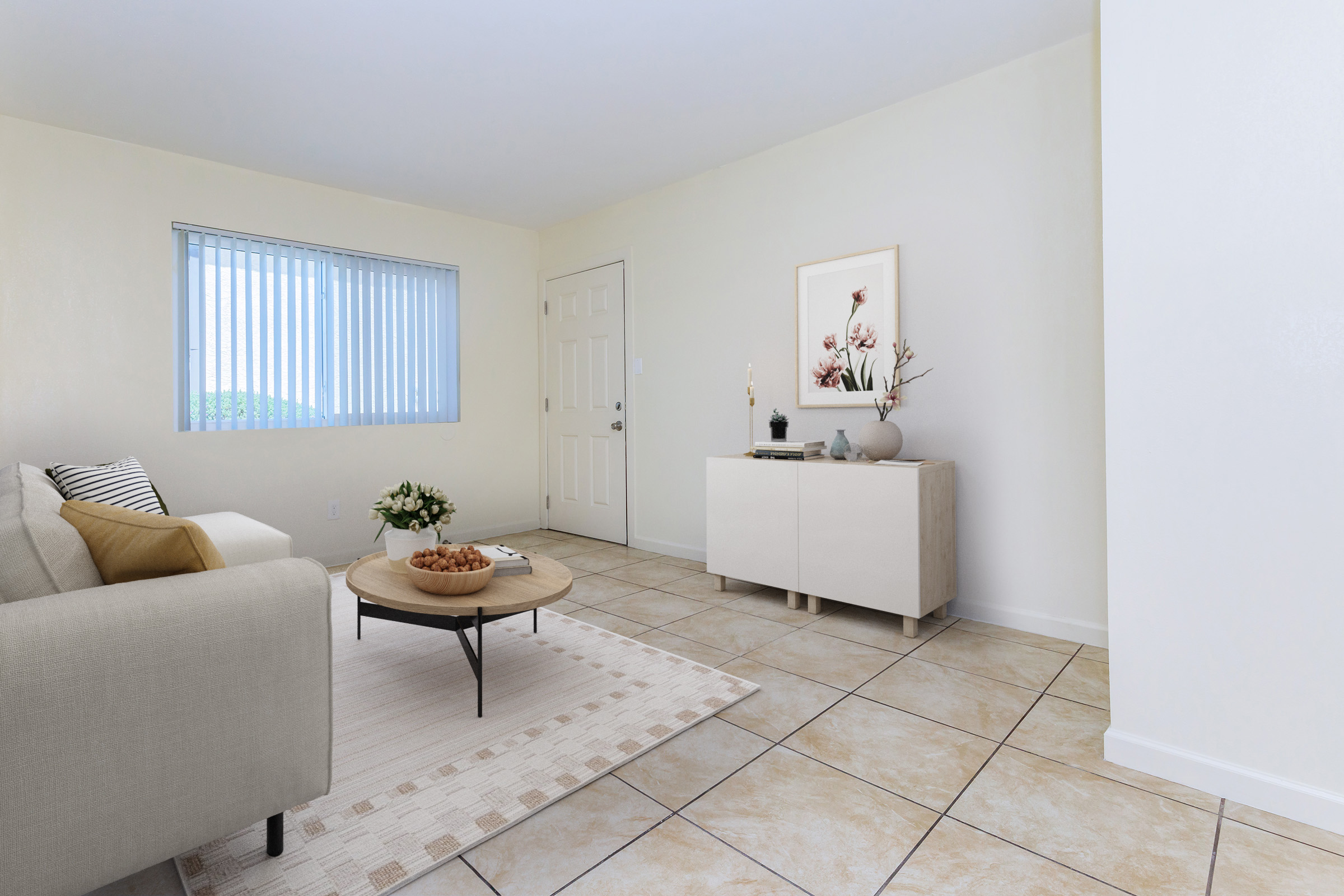 A bright, modern living room featuring a beige sofa with decorative cushions, a round coffee table with a bowl of fruit, and light-colored tiled flooring. A sideboard against a white wall holds decorative items, and a window with vertical blinds allows natural light to enter.