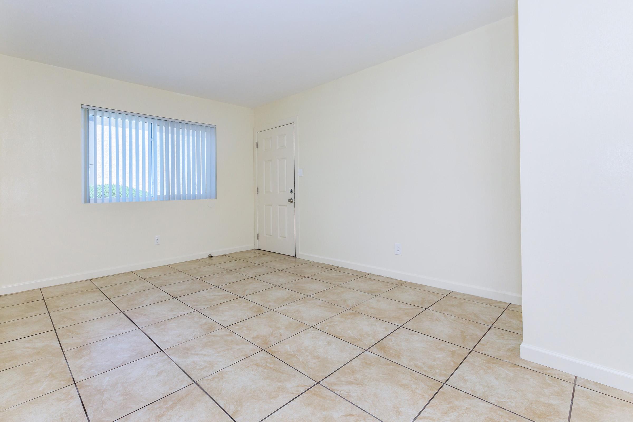 Empty room featuring light-colored walls, large windows with vertical blinds, and a tiled floor. The door is visible on one side, and the space is uncluttered, giving a sense of openness and potential for furnishings or decoration.