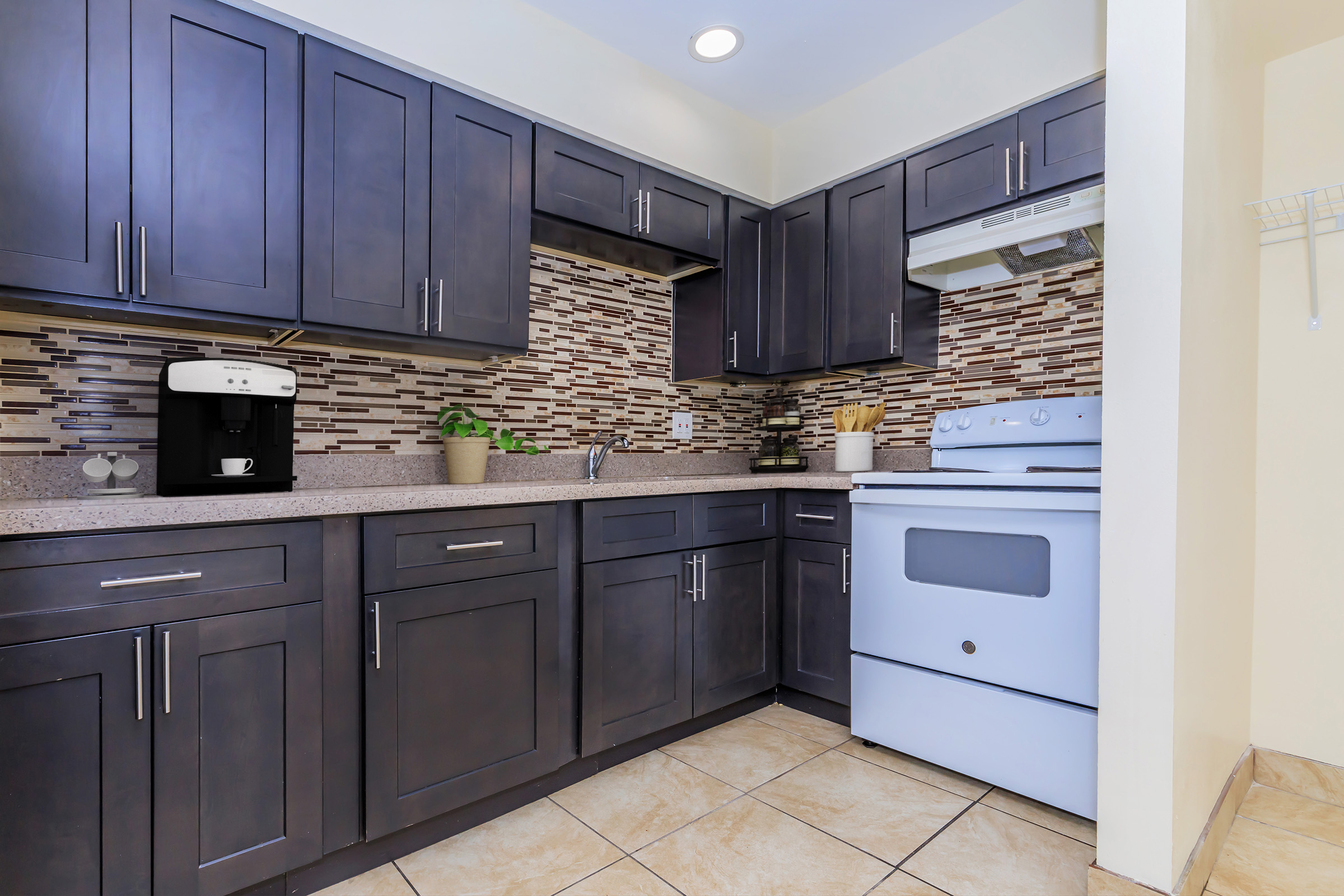 Modern kitchen featuring dark wood cabinets, a beige countertop, and a decorative backsplash made of small tiles. There's a white stove, a coffee maker, and a small plant on the counter. The floor is tiled, and the overall design is contemporary and inviting.
