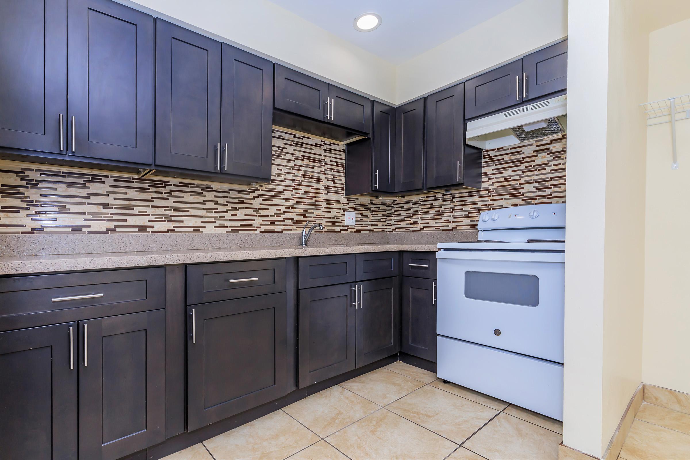 A modern kitchen featuring dark wood cabinets, a mosaic tile backsplash, and a white stove. The countertop is light-colored, and beige tiles cover the floor, creating a warm and inviting atmosphere.