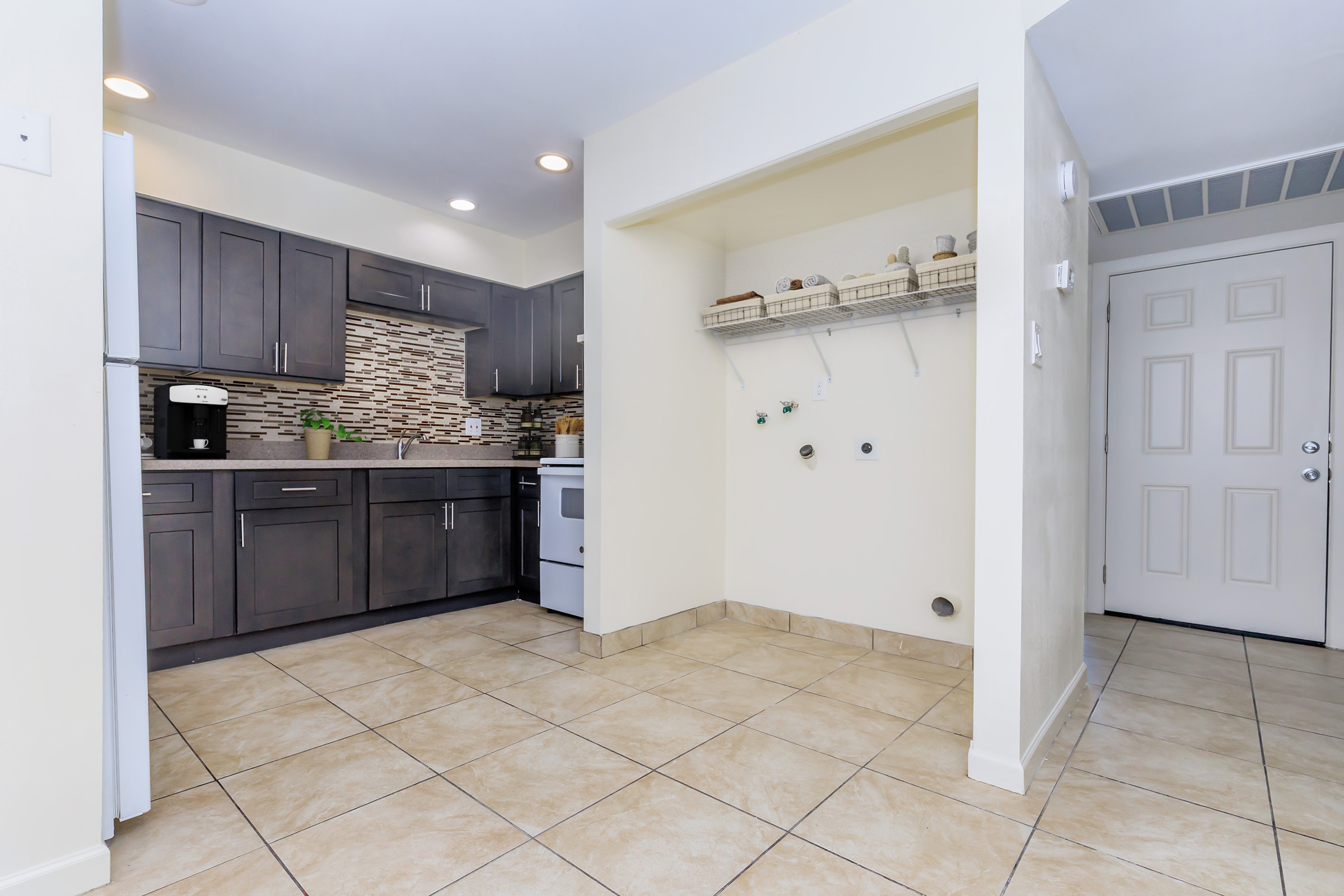 Modern kitchen featuring dark gray cabinets, a stone backsplash, and a white stove. The space has beige tiled flooring and a nearby wall with hooks and shelves. Natural light illuminates the area, creating a bright and inviting atmosphere. A door leads outside.