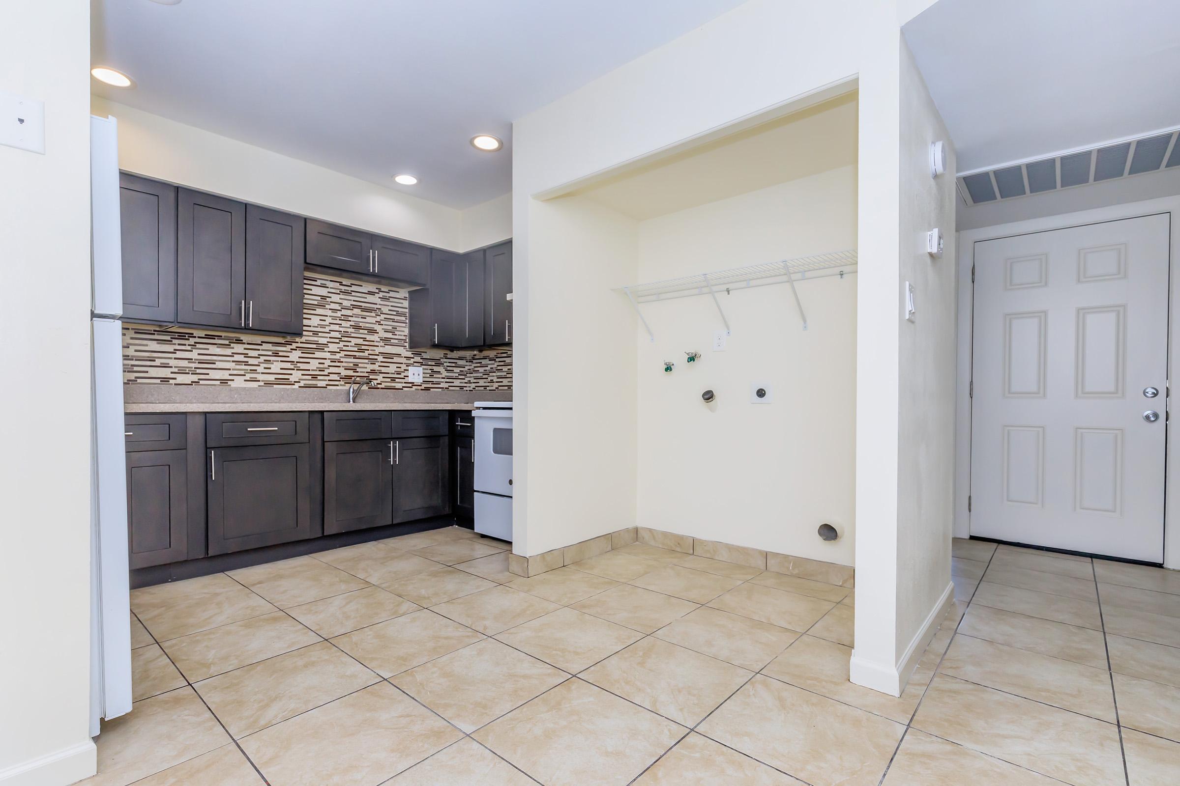 Modern kitchen with dark cabinets and a mosaic backsplash, featuring a white stove and refrigerator. The tile floor is light-colored, and there is an adjacent wall with a hook rack. A door leads out of the kitchen area, providing access to another room or hallway.