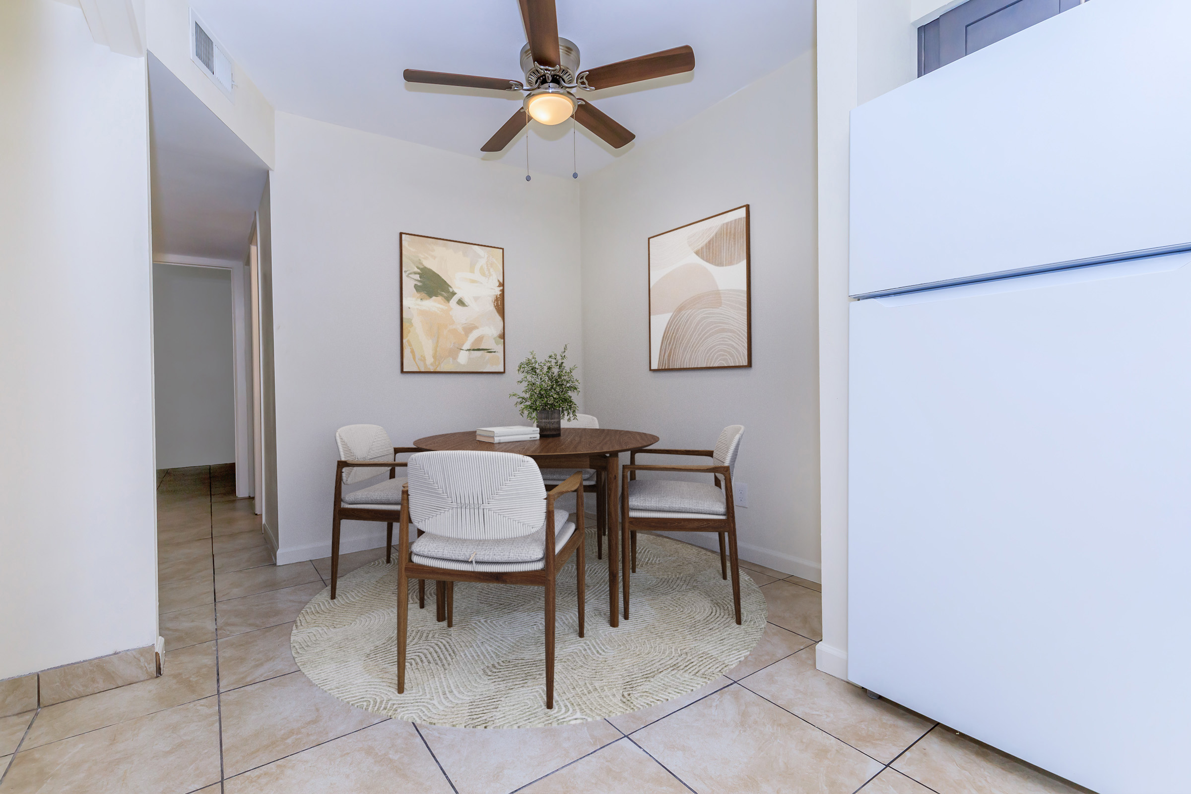 A cozy dining area featuring a round wooden table surrounded by four upholstered chairs. The space includes a white refrigerator, light-colored walls, and two framed artworks on the wall. A patterned area rug lies beneath the table, adding warmth to the room.