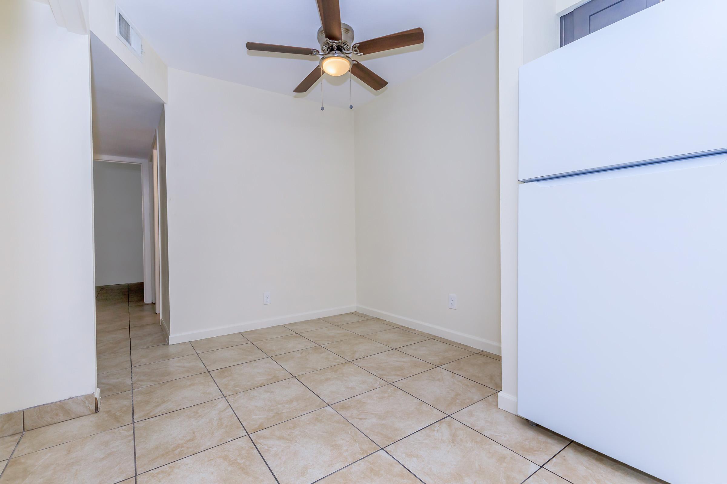 A simple, empty room featuring light-colored walls and tile flooring. A ceiling fan is mounted on the ceiling, providing a focal point. On one side, there is a white refrigerator, while the opposite side leads to a hallway. The space is well-lit, creating a clean and open atmosphere.