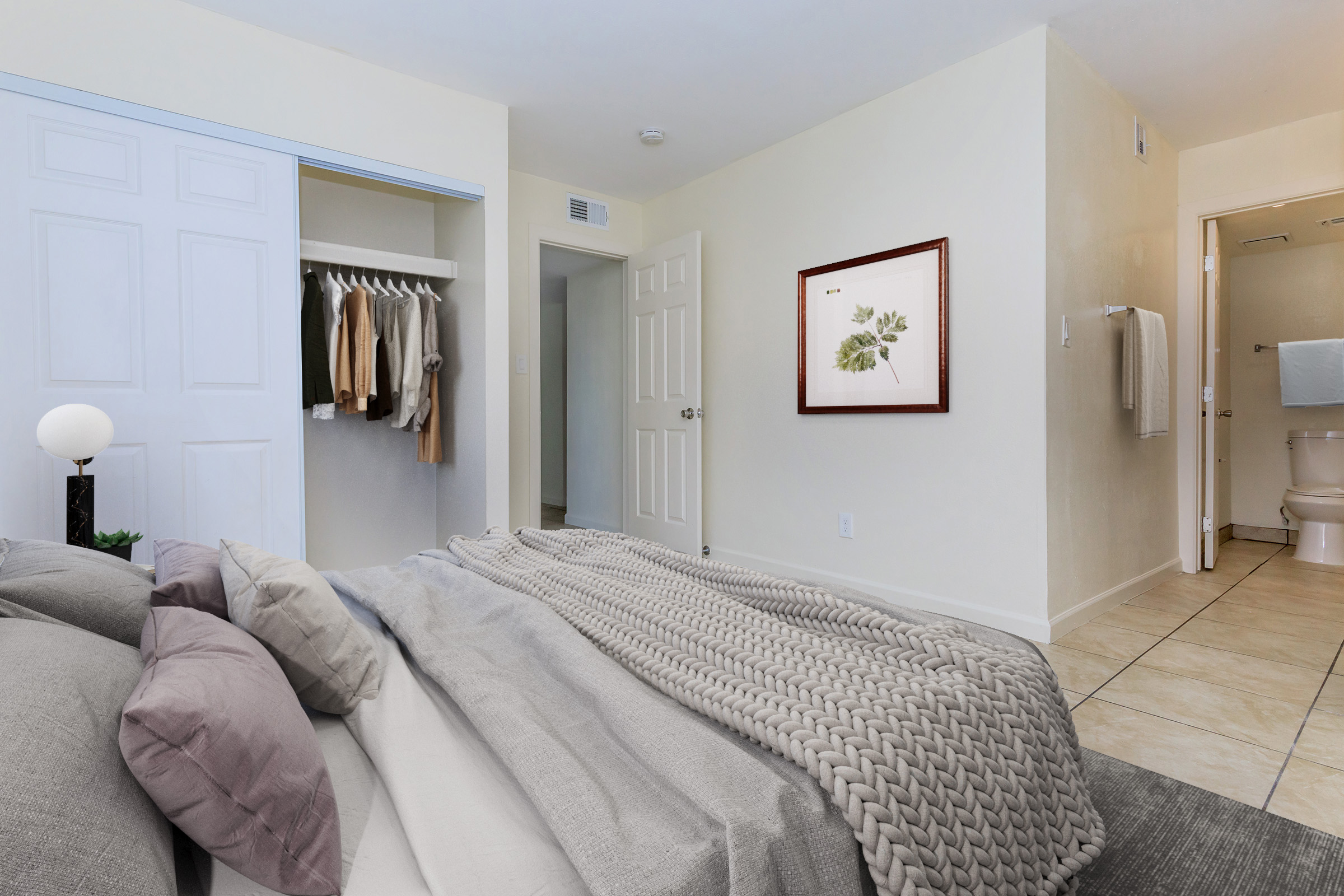 Cozy bedroom featuring a neatly made bed with gray and taupe bedding, a textured throw blanket, and a bedside lamp. A closet with hanging clothes is visible, along with a doorway leading to a bathroom. The room has light-colored walls and ceramic tile flooring, creating a warm and inviting atmosphere.