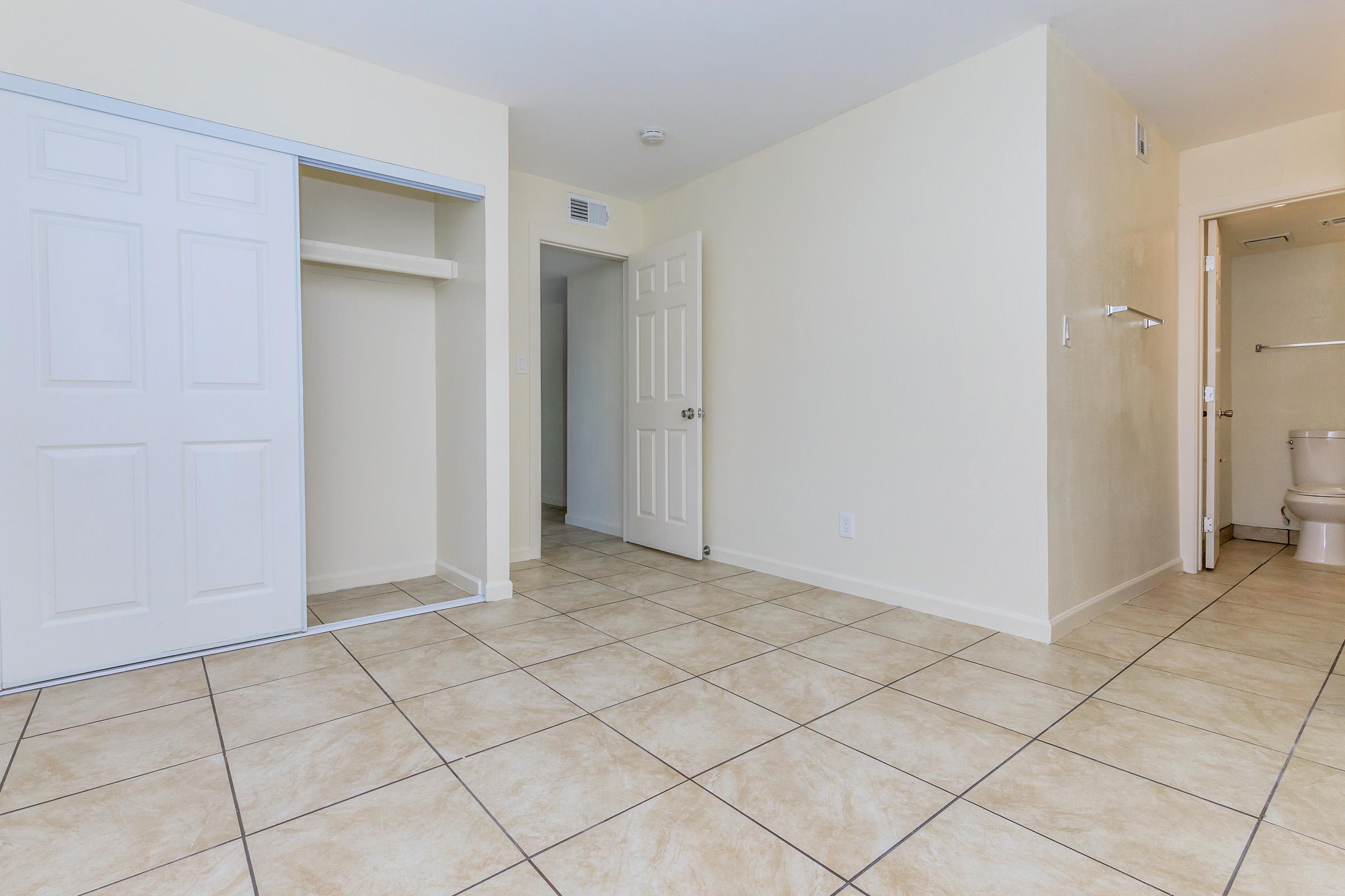 A spacious, empty room with beige walls and tiled flooring. There's a closet with sliding doors on the left, a doorway leading to another room ahead, and a bathroom door on the right. The overall ambiance is bright and inviting, with ample natural light.