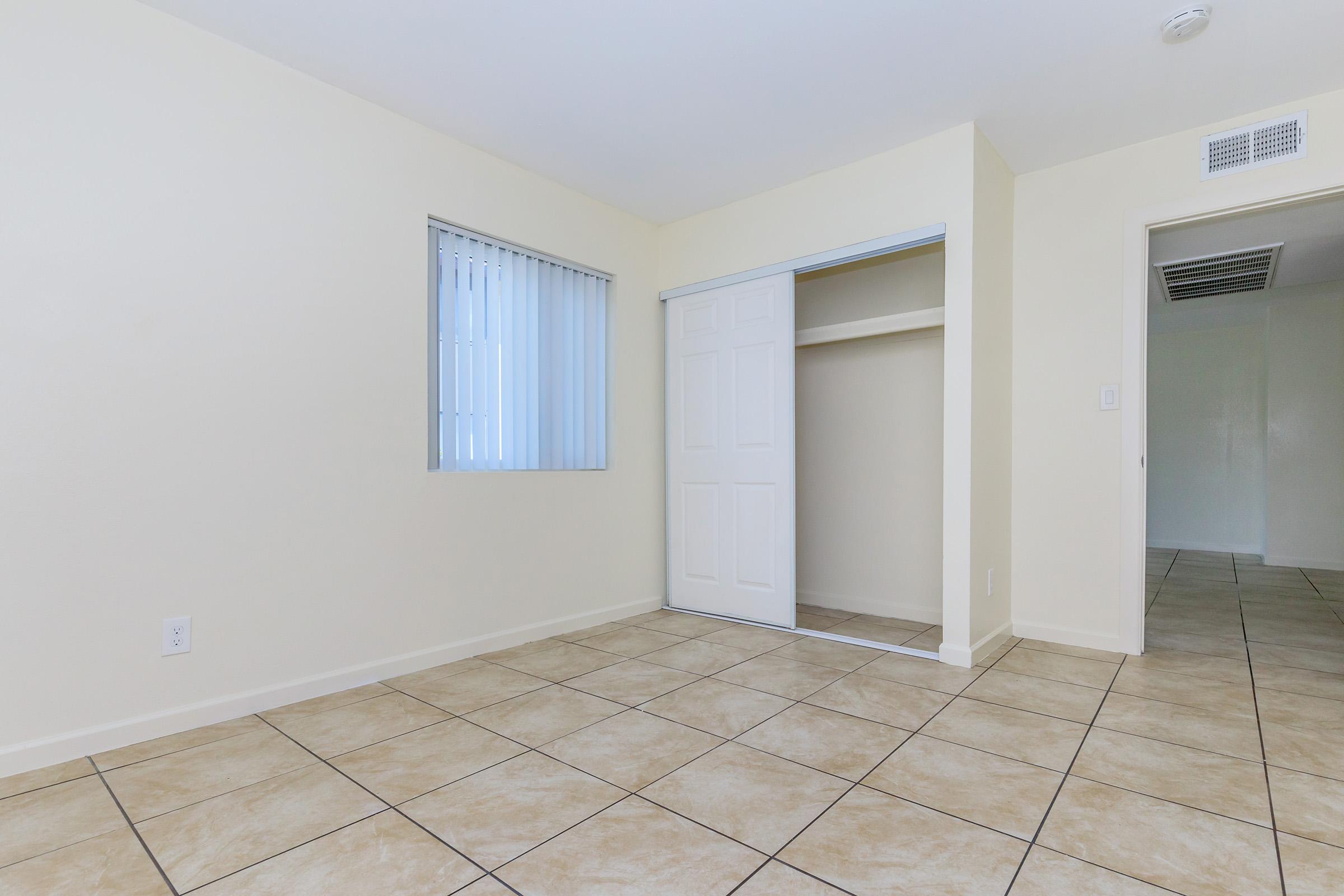 A bright and empty room with light-colored walls and tiled flooring. One wall features a vertical window with blinds, while another wall has a closet with sliding doors. The room has a simple, clean design, creating a spacious and airy feel. A doorway leads to another room in the background.