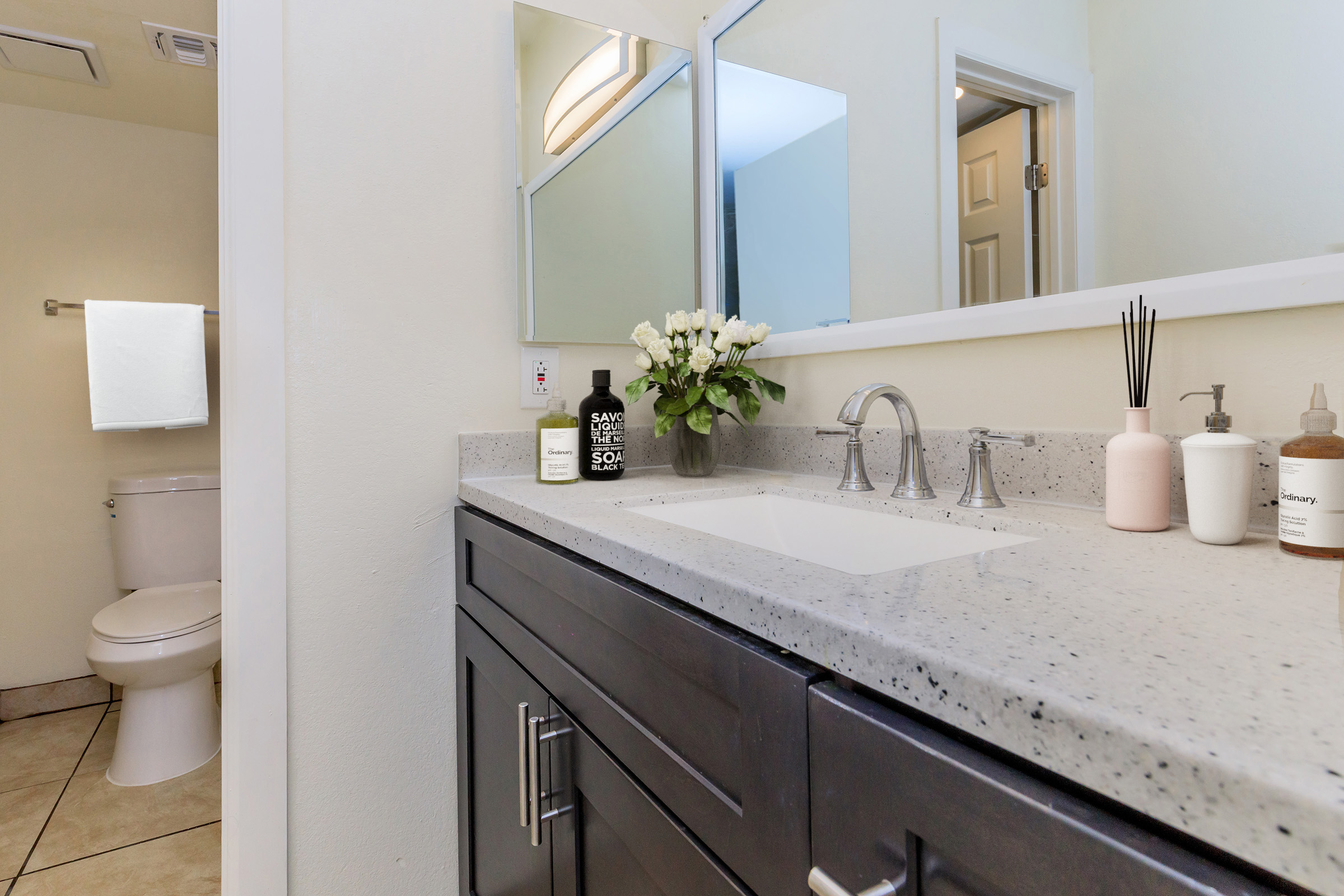 A modern bathroom featuring a gray vanity with a white countertop, a vase of flowers, and various toiletries. In the background, a toilet is partially visible, along with a towel hanging on the wall. Natural light enters through a nearby window, enhancing the clean and tidy appearance of the space.
