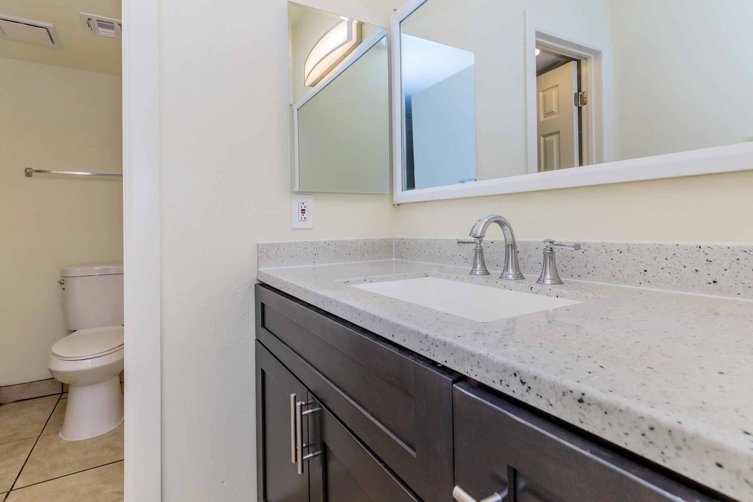 A modern bathroom featuring a sleek countertop with a double sink, a large mirror above, and a light-colored wall. In the background, there is a white toilet and a towel rack. The floor is tiled, adding to the contemporary design.