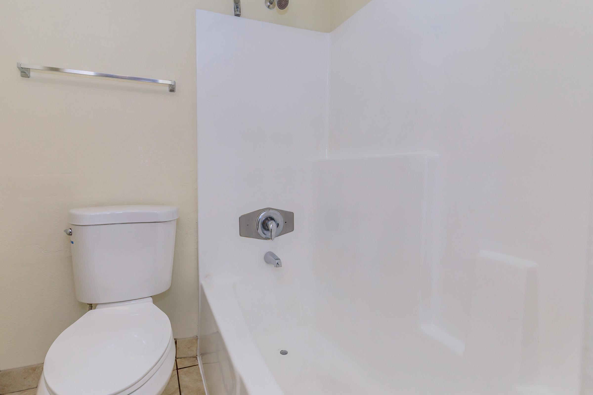 A clean bathroom featuring a white bathtub and a toilet. The bathtub is adjacent to a wall and has a faucet and showerhead. The bathroom has light-colored tiles on the floor and a simple towel rack mounted on the wall. The overall appearance is neat and minimalistic.