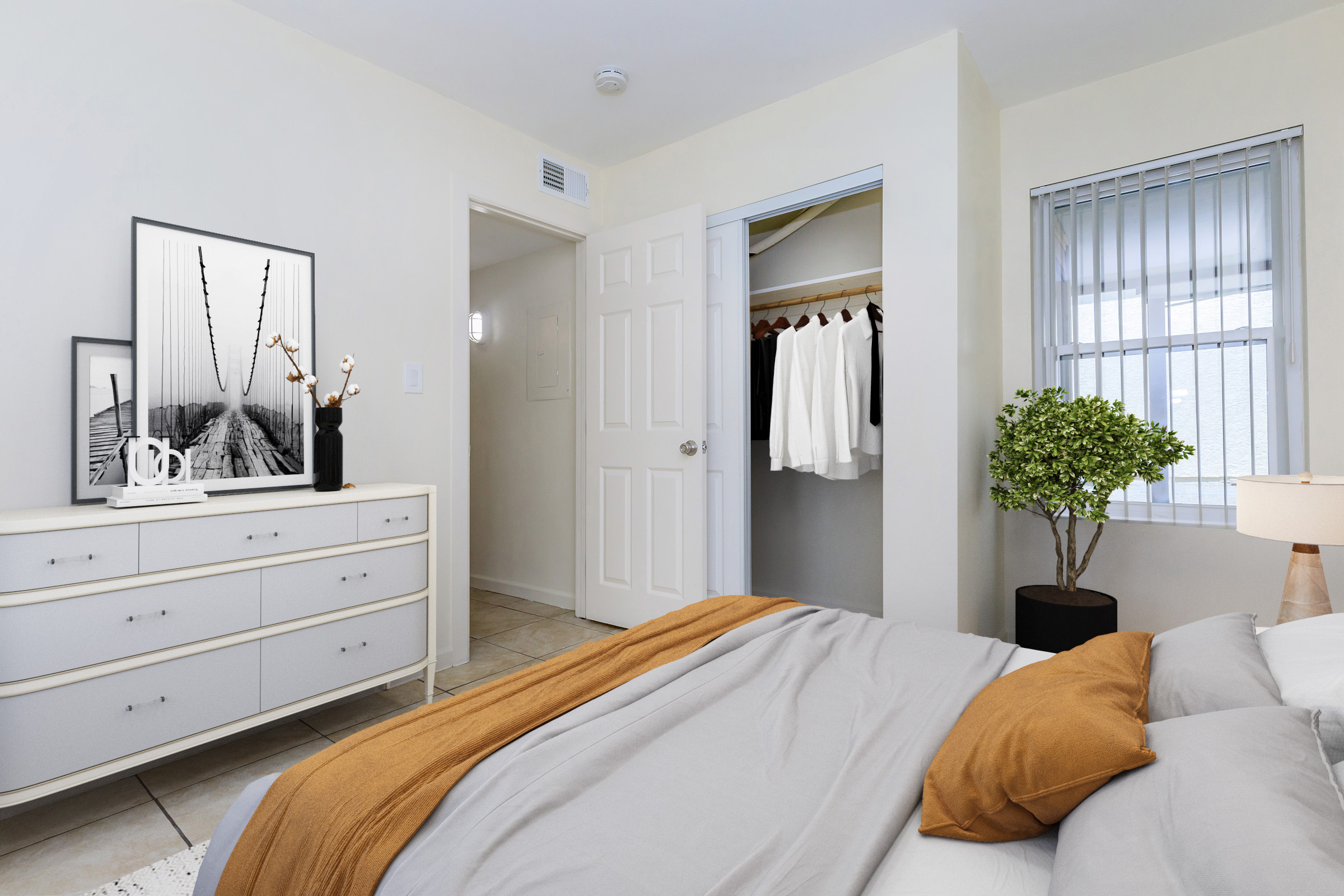 A neatly arranged bedroom featuring a light color palette. There is a bed with gray and brown bedding, a dresser with a framed black-and-white photograph, a small potted plant, and a lamp. A closet is seen with hanging clothes, and natural light is coming through a window with blinds. The room has a clean, modern aesthetic.