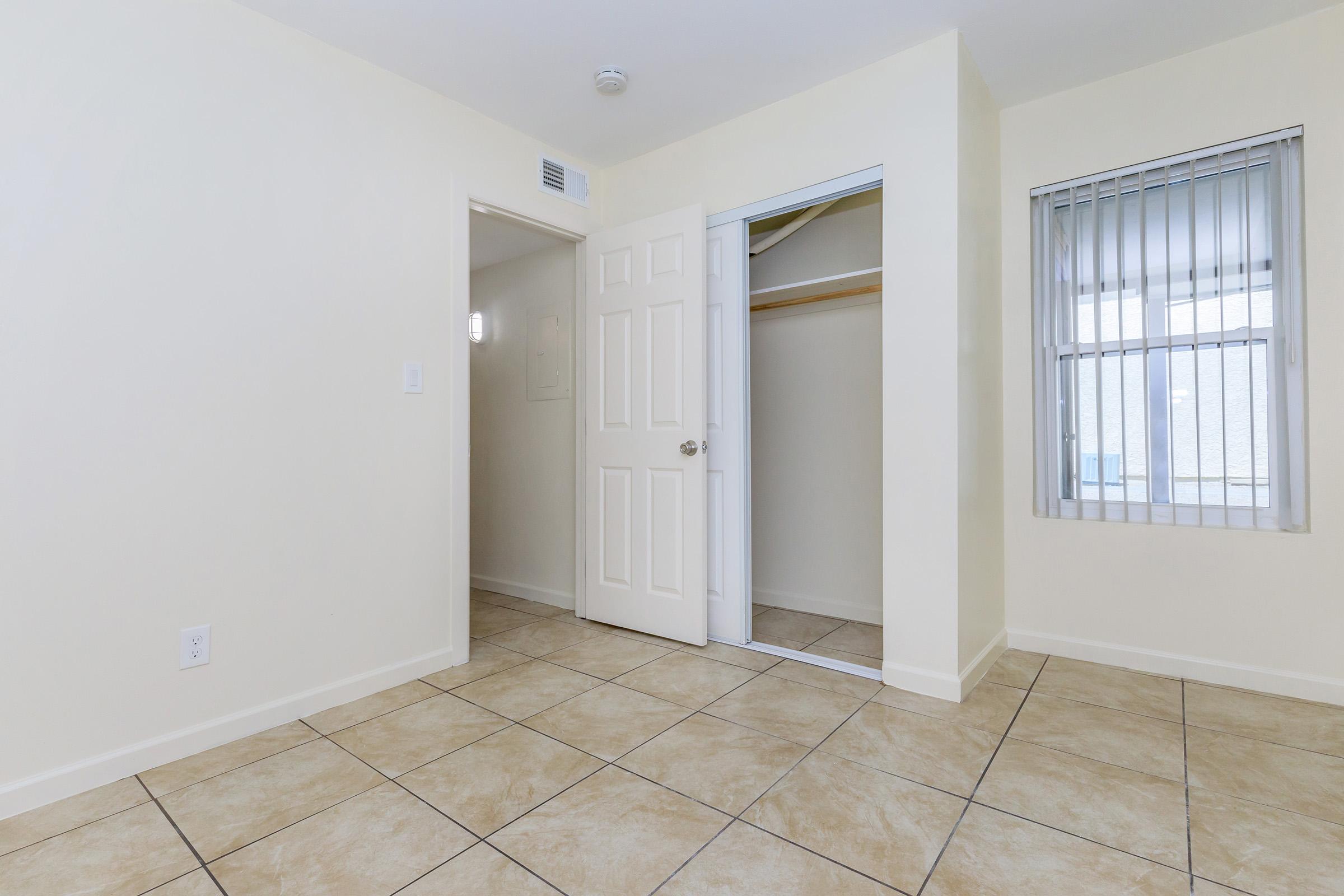 A light-colored room featuring beige tiled flooring, a closed white door leading to another space, and an open closet with a hanging rod. A window with blinds allows natural light, brightening the room. The walls are painted in a soft hue, creating a clean and minimalist atmosphere.