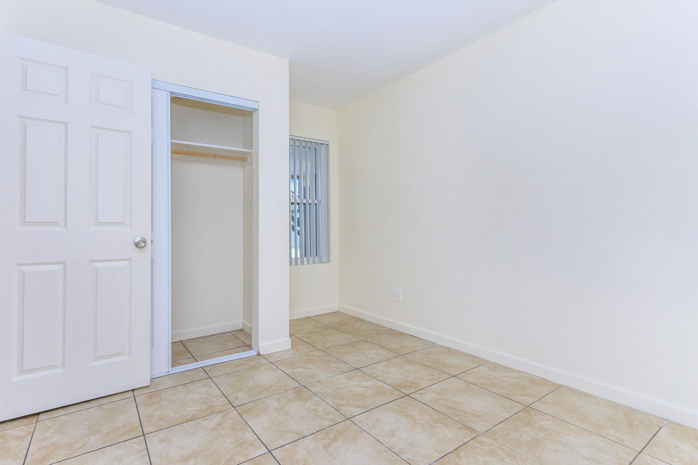Empty room featuring beige walls, tiled flooring, and a closet with a closed door. A window with vertical blinds allows natural light. The space is minimalistic and unadorned, suitable for various uses or decoration.