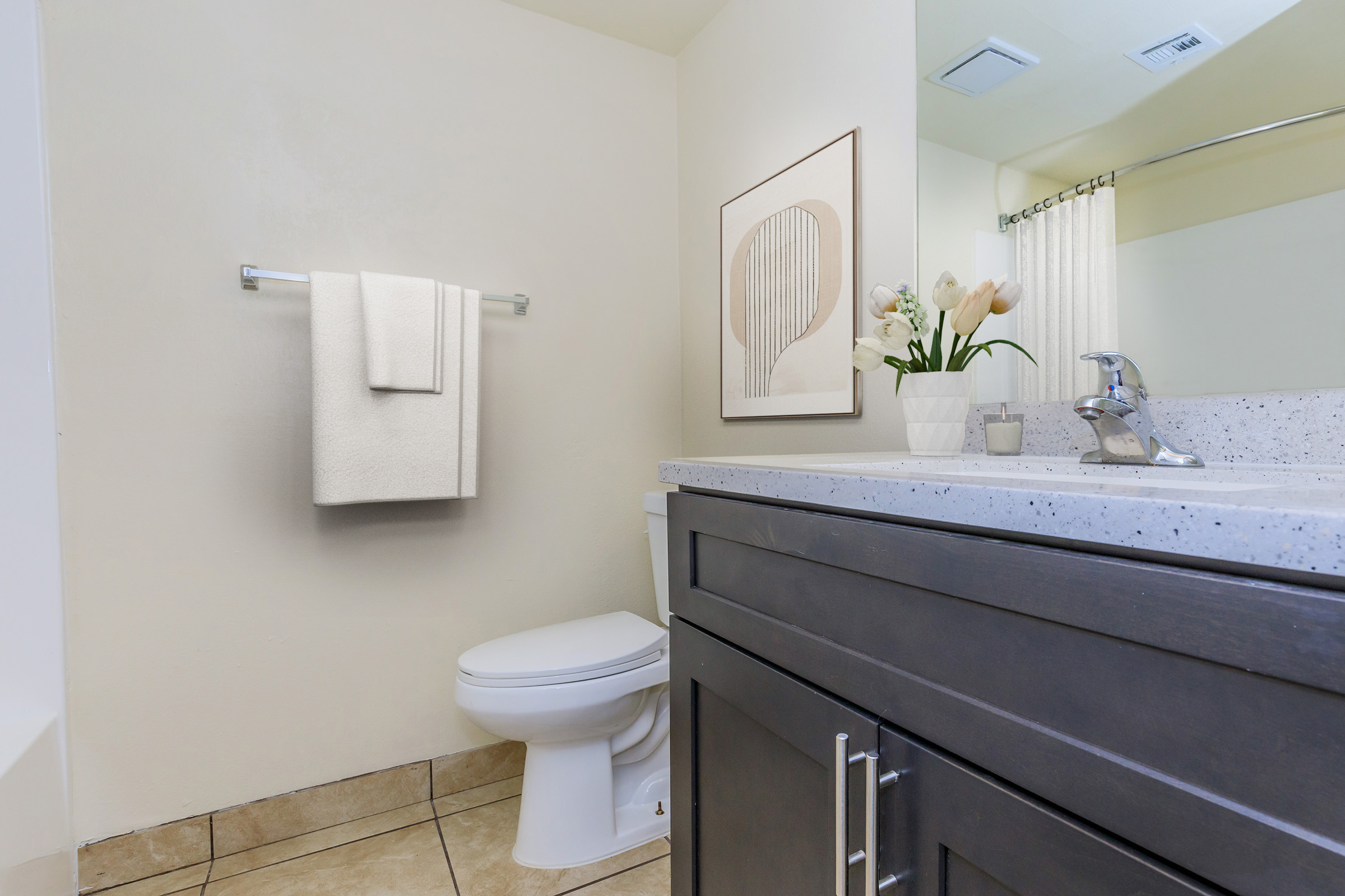 A modern bathroom featuring a gray vanity with a light-colored countertop, a white toilet, and neatly hung towels. A small potted plant and artwork are displayed on the countertop, with natural light illuminating the space through a mirror and window. The tiled floor adds to the contemporary design.
