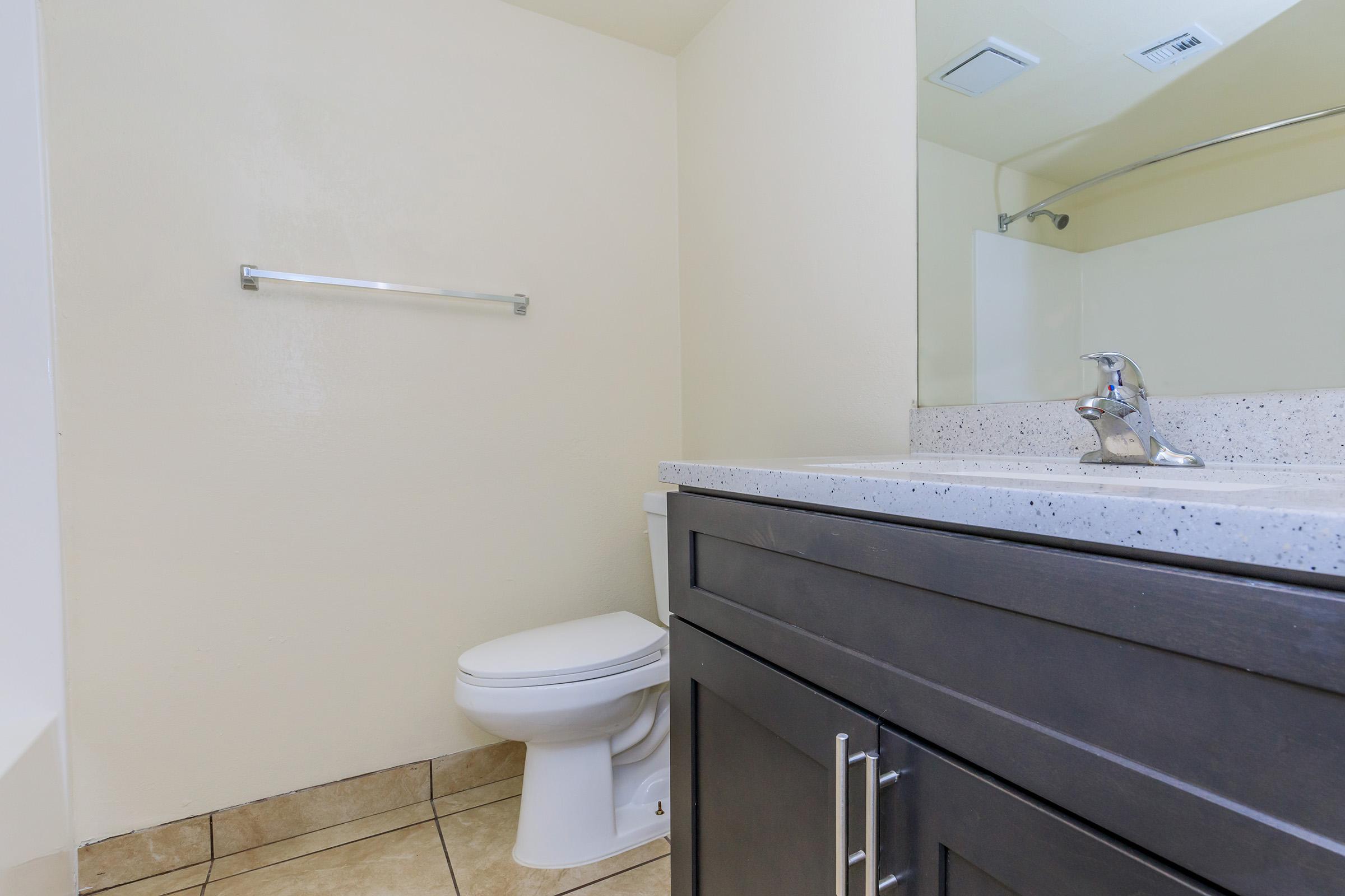 A clean and modern bathroom featuring a toilet, a sink with a chrome faucet on a speckled countertop, and a large mirror. The walls are painted a light color, and there is a towel bar mounted above the toilet. The flooring consists of tiled surfaces, contributing to a fresh and tidy appearance.