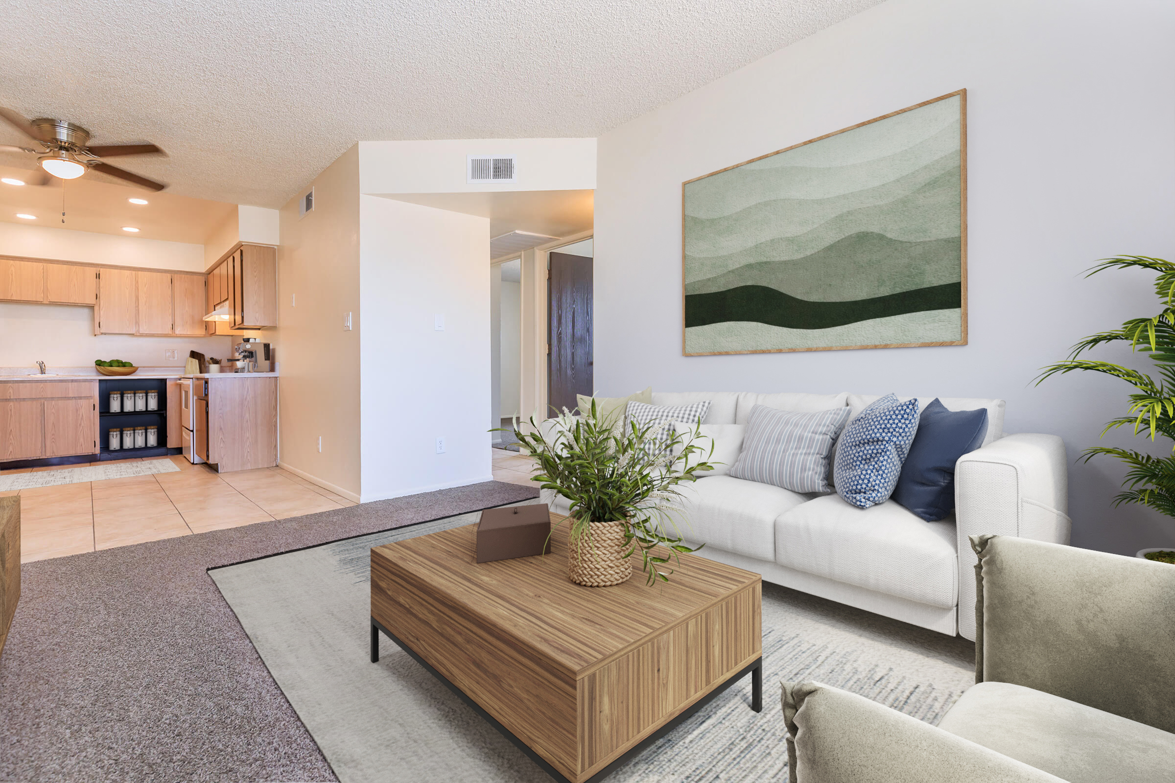 A modern living room featuring a white sofa with blue and gray decorative pillows, a wooden coffee table, and a large green abstract art piece on the wall. There is a potted plant in the corner, and the open kitchen can be seen in the background with wood cabinets and countertops. The flooring is a mix of tile and carpet.