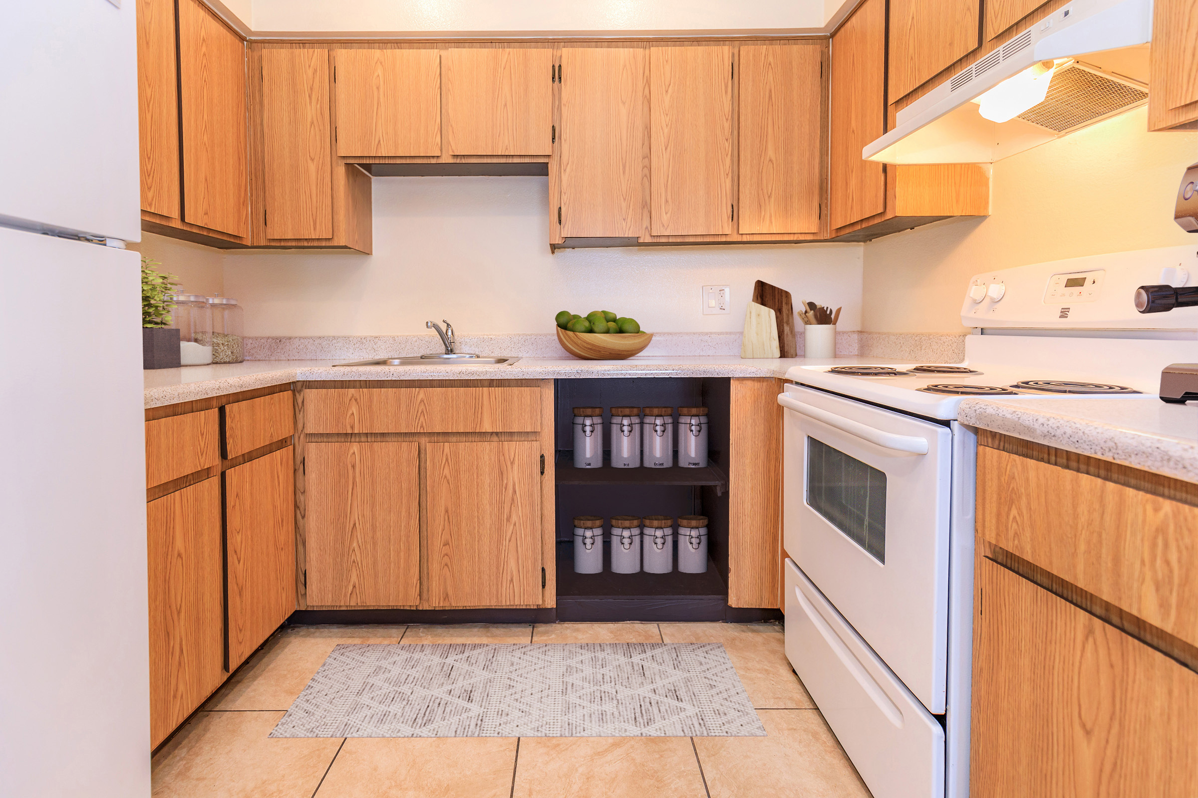 A modern kitchen featuring wooden cabinets, a countertop with a bowl of green apples, and a stove. The space includes an organized area with glass containers on a lower shelf, beige tile flooring, and a soft area rug. Natural light illuminates the room, creating a warm and inviting atmosphere.