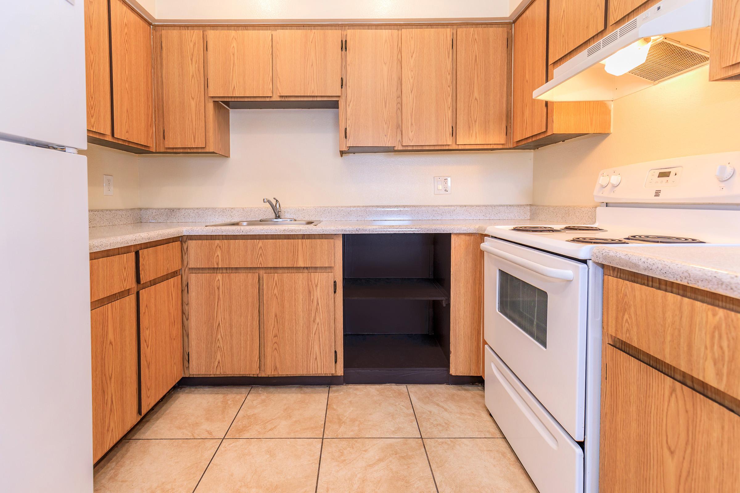 A kitchen featuring light wood cabinets, a white stove, and a refrigerator. The countertop is beige with a single sink. The tile floor is light-colored. There is an open space under the counter, and the walls are painted a soft yellow.