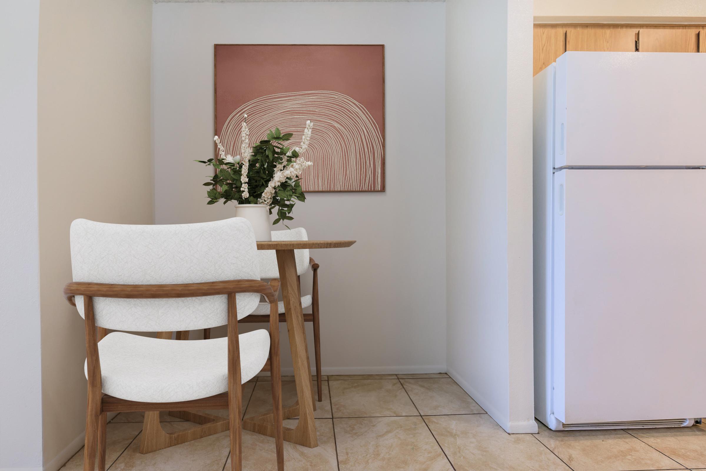 A small dining area featuring a wooden table with two white upholstered chairs. A simple vase with greenery sits on the table. The wall above has a minimalist art piece with curved lines in warm tones. To the side, there is a white refrigerator, and the floor is tiled in light colors.