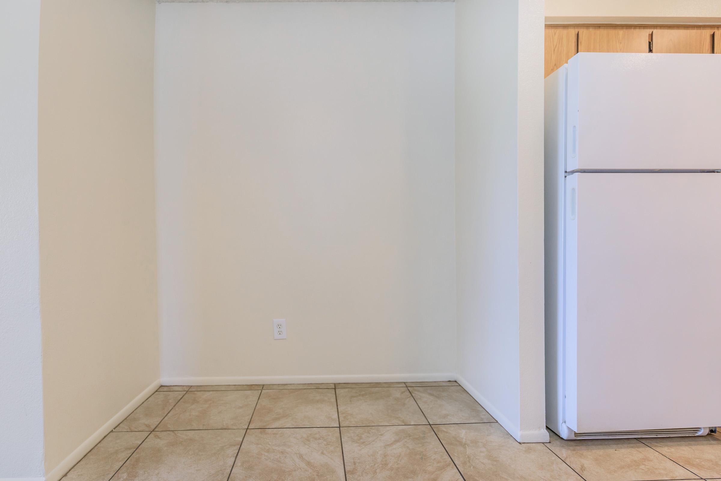 Empty kitchen space with beige tiled floor, light-colored walls, and a white refrigerator partially visible on the right. The area is uncluttered, featuring no furniture or decorations, providing a blank canvas for future use.