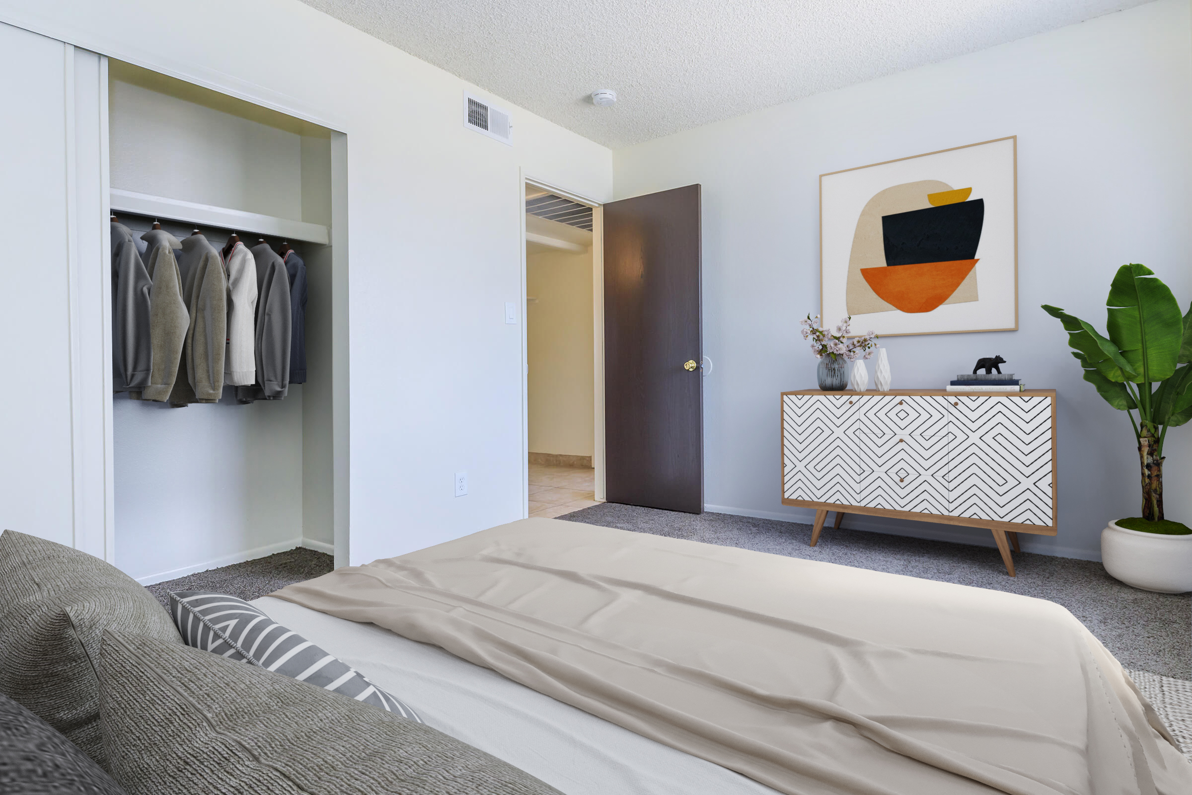 A modern bedroom featuring a neatly made bed with a light beige blanket, a stylish dresser with geometric patterns, a closet with hanging clothes, a large abstract wall art, and a potted plant in the corner. The room has neutral-colored walls and carpet, with a door leading to another space.