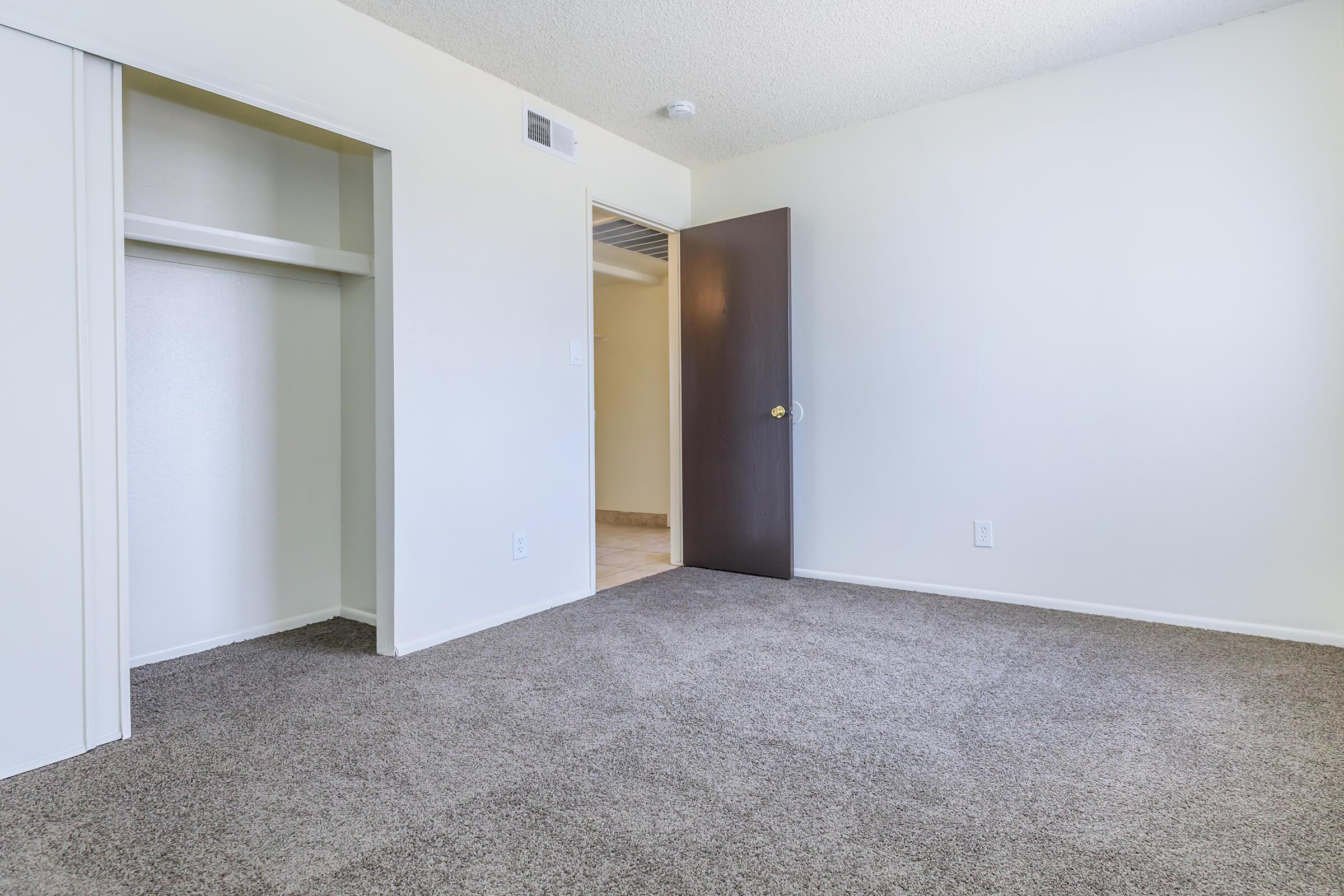 A vacant room featuring light-colored walls and gray carpet. There is a closet on the left and a closed door on the right leading to another area. The room has natural light and is unfurnished, giving a sense of space and potential for personalization.