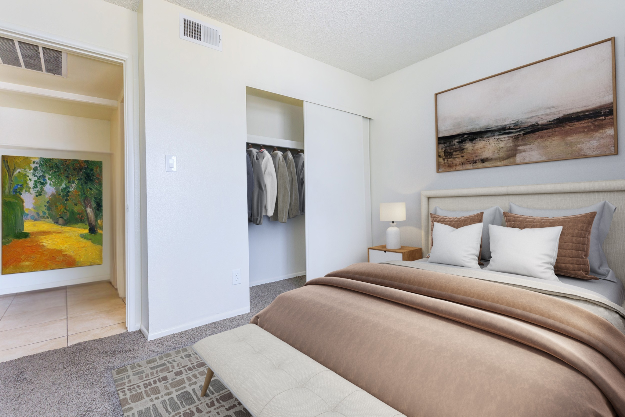 A warmly decorated bedroom featuring a neatly made bed with brown and gray bedding. A bedside lamp casts a soft glow. In the background, a closet with hanging clothing is visible, alongside a hallway leading to a colorful painting of a landscape. The overall ambiance is cozy and inviting.