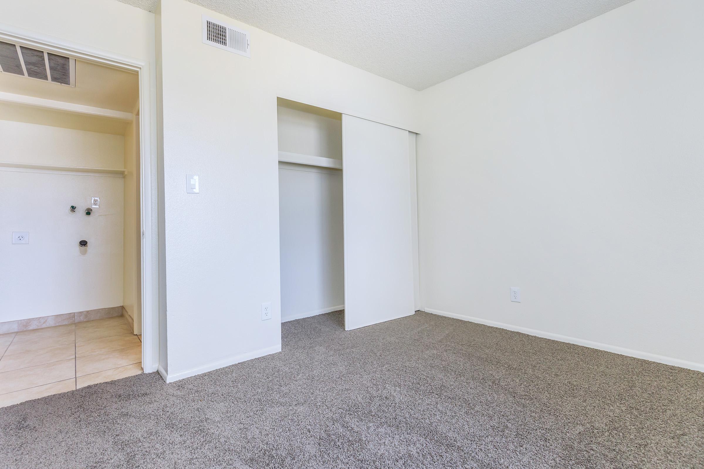 A spacious, empty room with light-colored walls and carpet. A closet is visible with sliding doors on one side, and an adjacent wall features a small utility area with plumbing. The floor is tiled near the entrance, suggesting a functional living space.