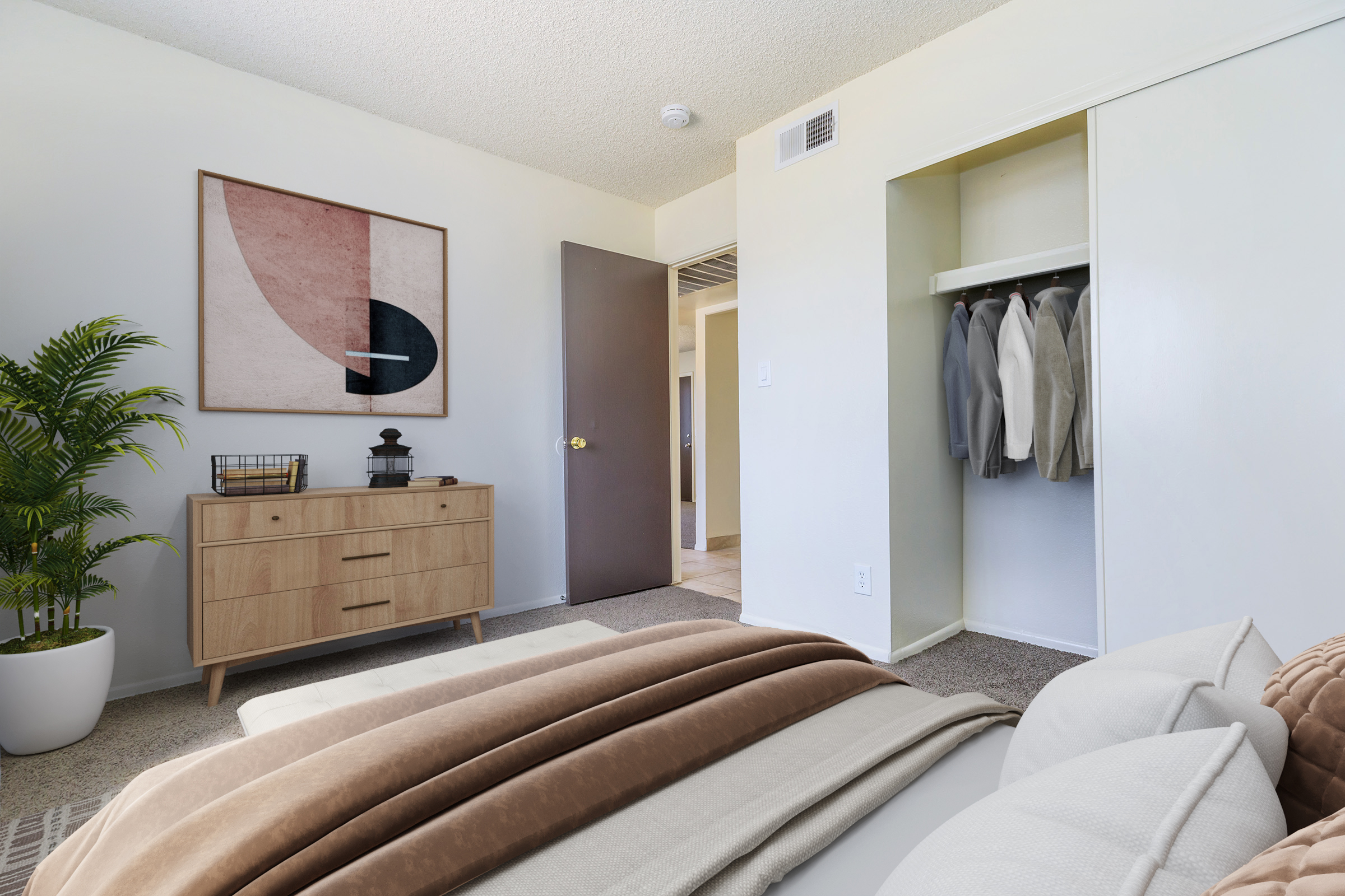 A cozy bedroom featuring a neatly made bed with neutral-toned bedding, a wooden dresser with a modern art piece above it, a potted plant in the corner, and a closet with neatly hung garments. The wall color is light, and there’s a door leading to another room in the background. Natural light brightens the space.