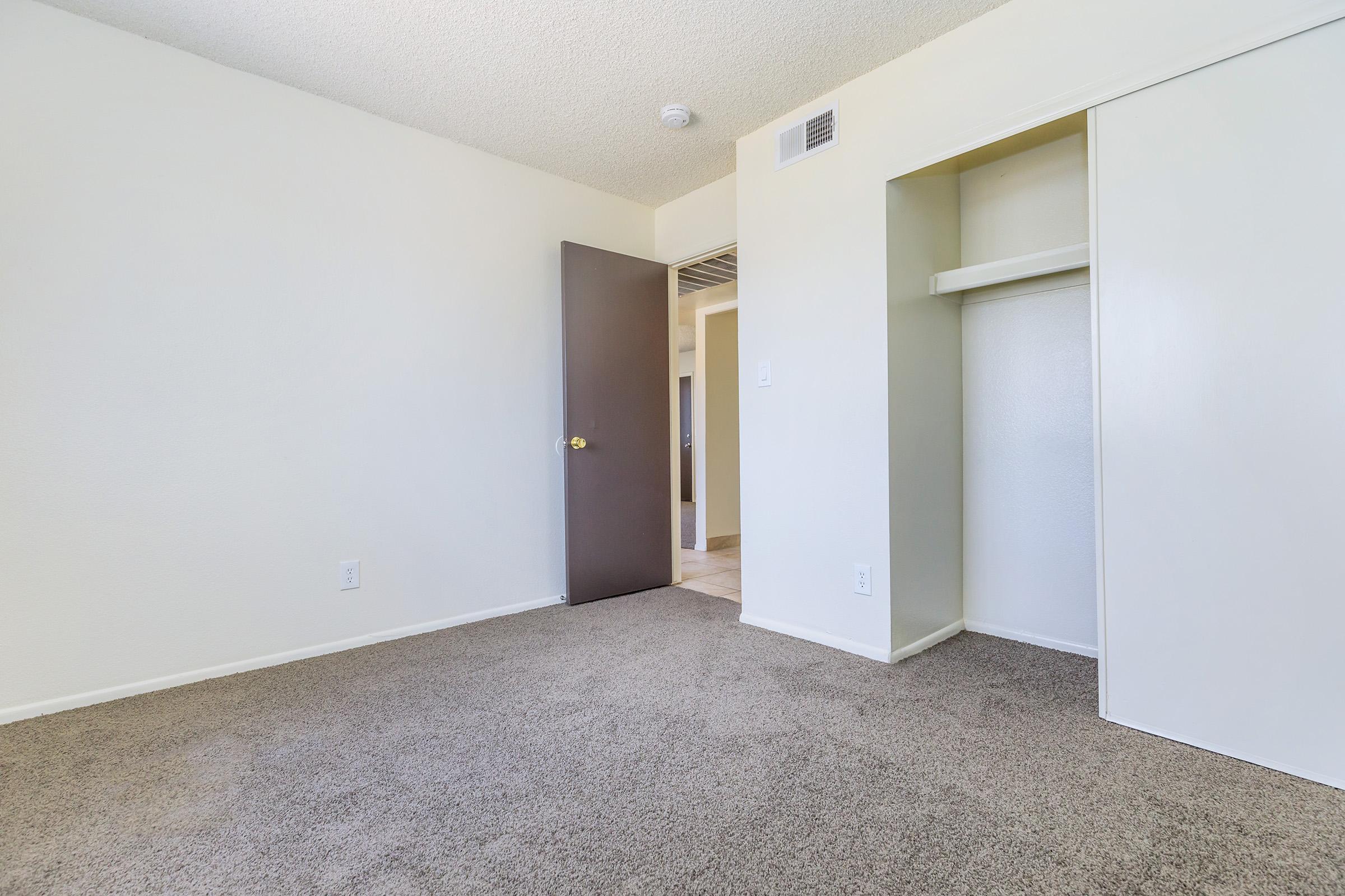 A light-colored room with beige carpet, featuring a closed dark brown door leading to another space. On the right, there is a small closet with an open shelf, and the walls are painted a soft off-white. Natural light enters from an unseen window, highlighting the spacious feel of the room.