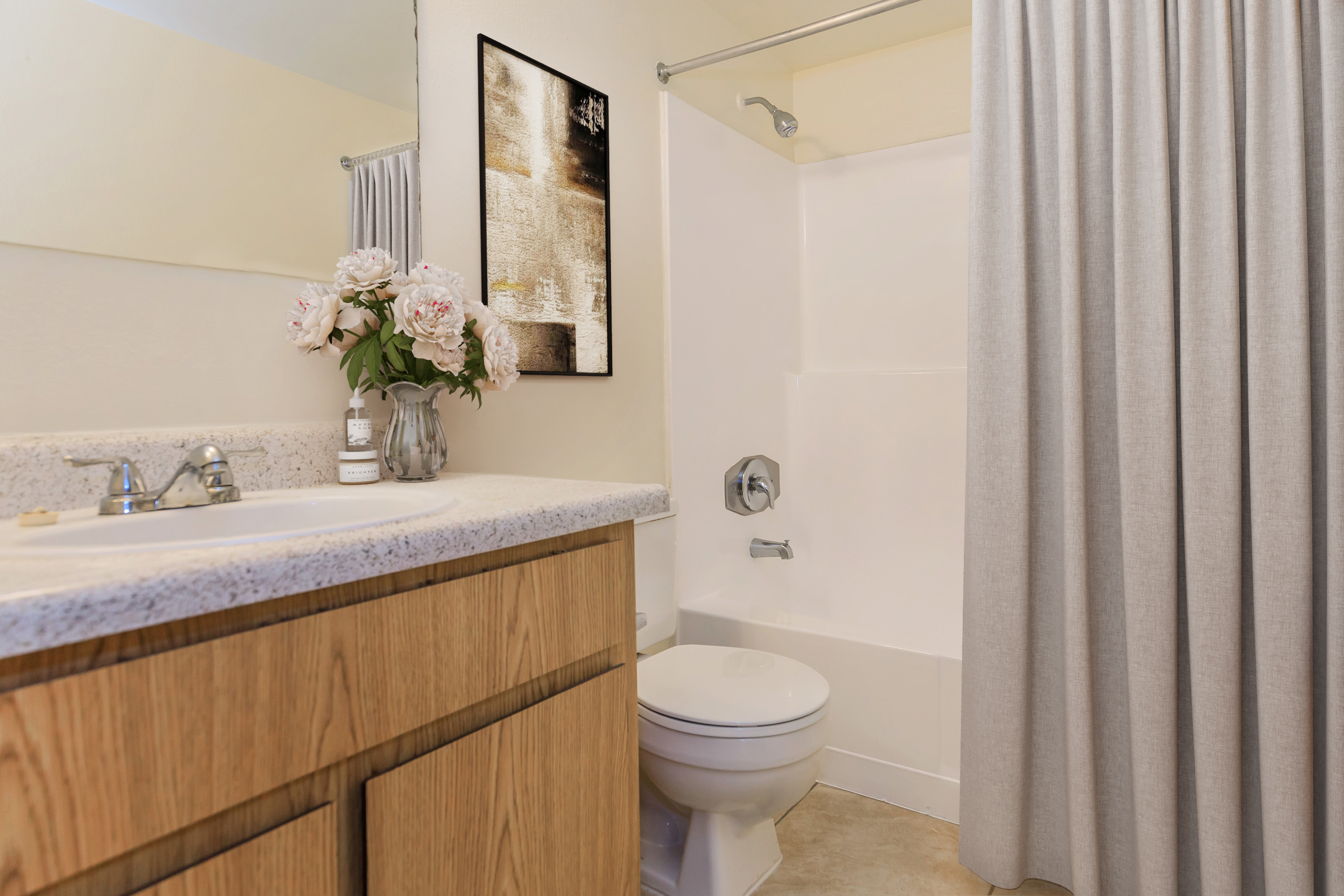 A clean bathroom featuring a countertop with a vase of pink peonies, a sink with chrome faucets, and a white toilet. The shower area is enclosed with a grey curtain, and there's a modern piece of art on the wall, creating a serene and inviting atmosphere.