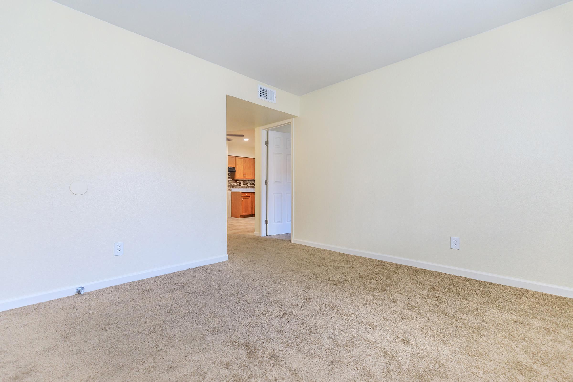 A spacious, empty room with light-colored walls and a beige carpeted floor. A doorway leads to another room, suggesting an open floor plan. The ambient lighting is bright, creating a welcoming atmosphere.