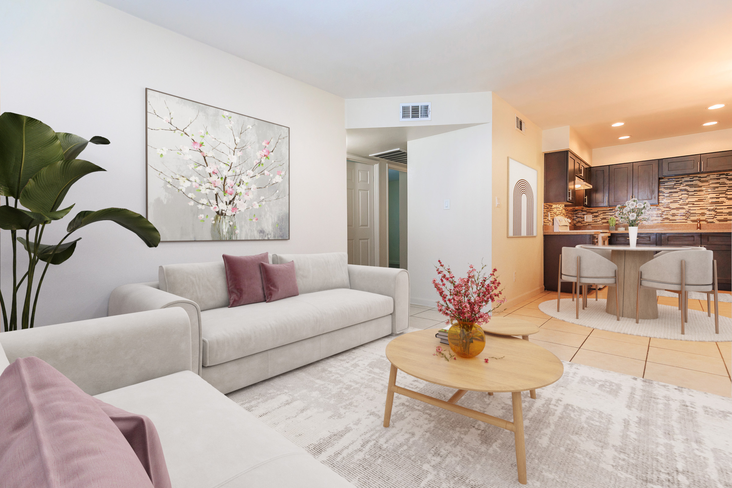 A modern living room featuring a light-colored sofa with pink cushions, a round wooden coffee table with a vase of flowers, and a wall art piece depicting a flowering branch. There's a potted plant in the corner, and the adjoining kitchen area is visible in the background, showcasing dark cabinetry and decorative backsplash.