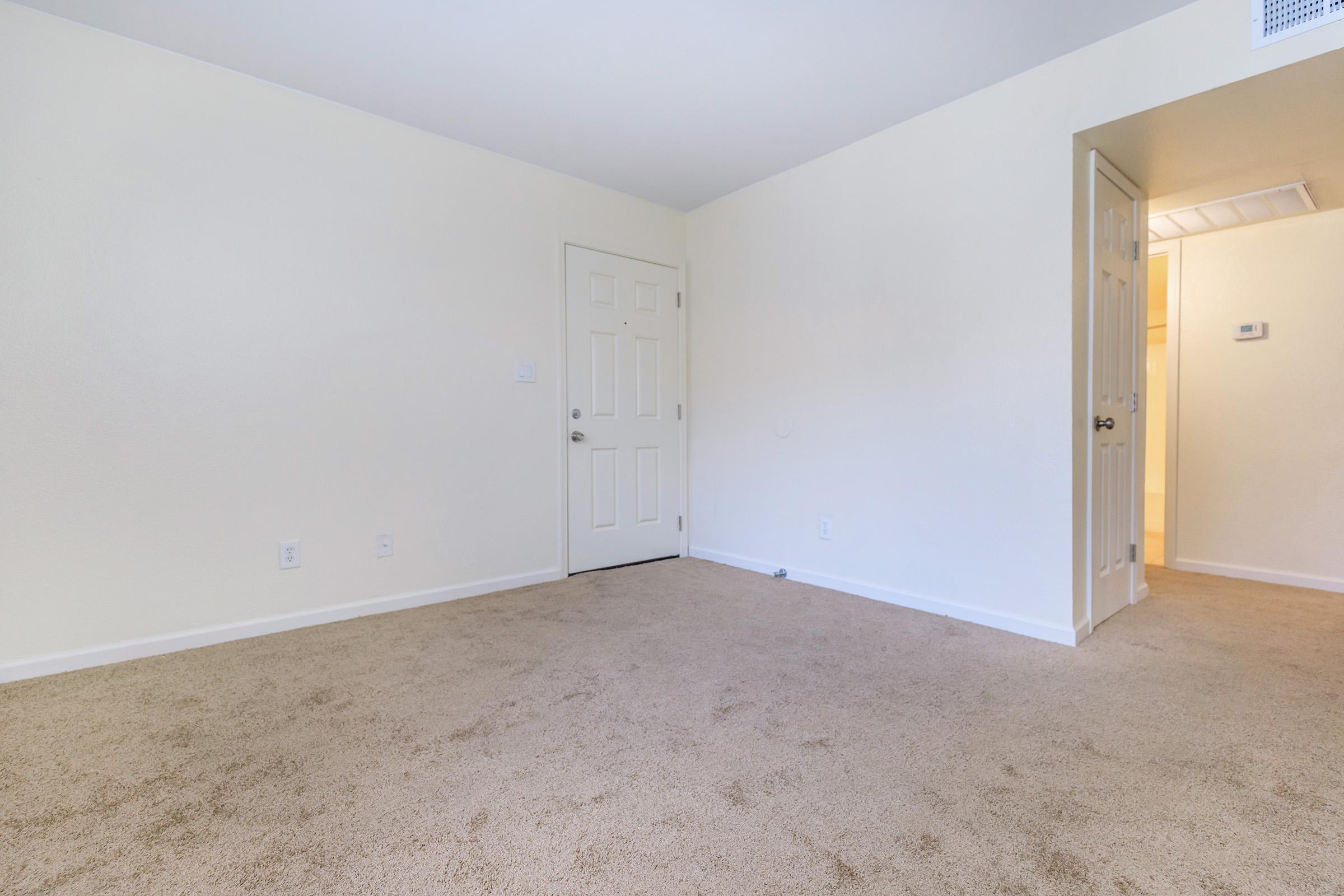 A spacious, empty room with light beige carpet and white walls. There is a closed door on the left side of the image and a doorway on the right leading to a hallway with warm lighting. The room appears clean and modern, ideal for various purposes such as a living area or office space.