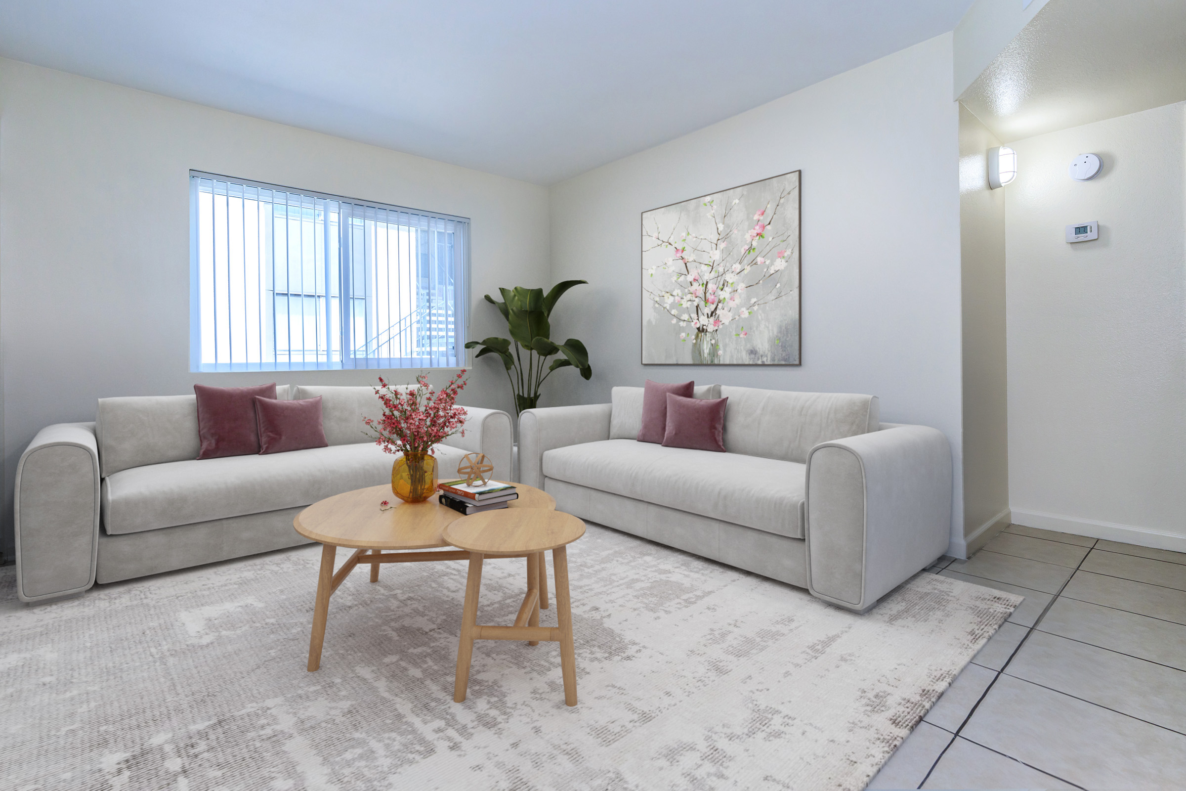 A modern living room featuring two light gray sofas with pink cushions facing a round wooden coffee table. A vase of flowers sits on the table, and a piece of artwork with floral designs decorates the wall. A window provides natural light, and there’s a potted plant in the corner. Neutral colors and a cozy atmosphere.
