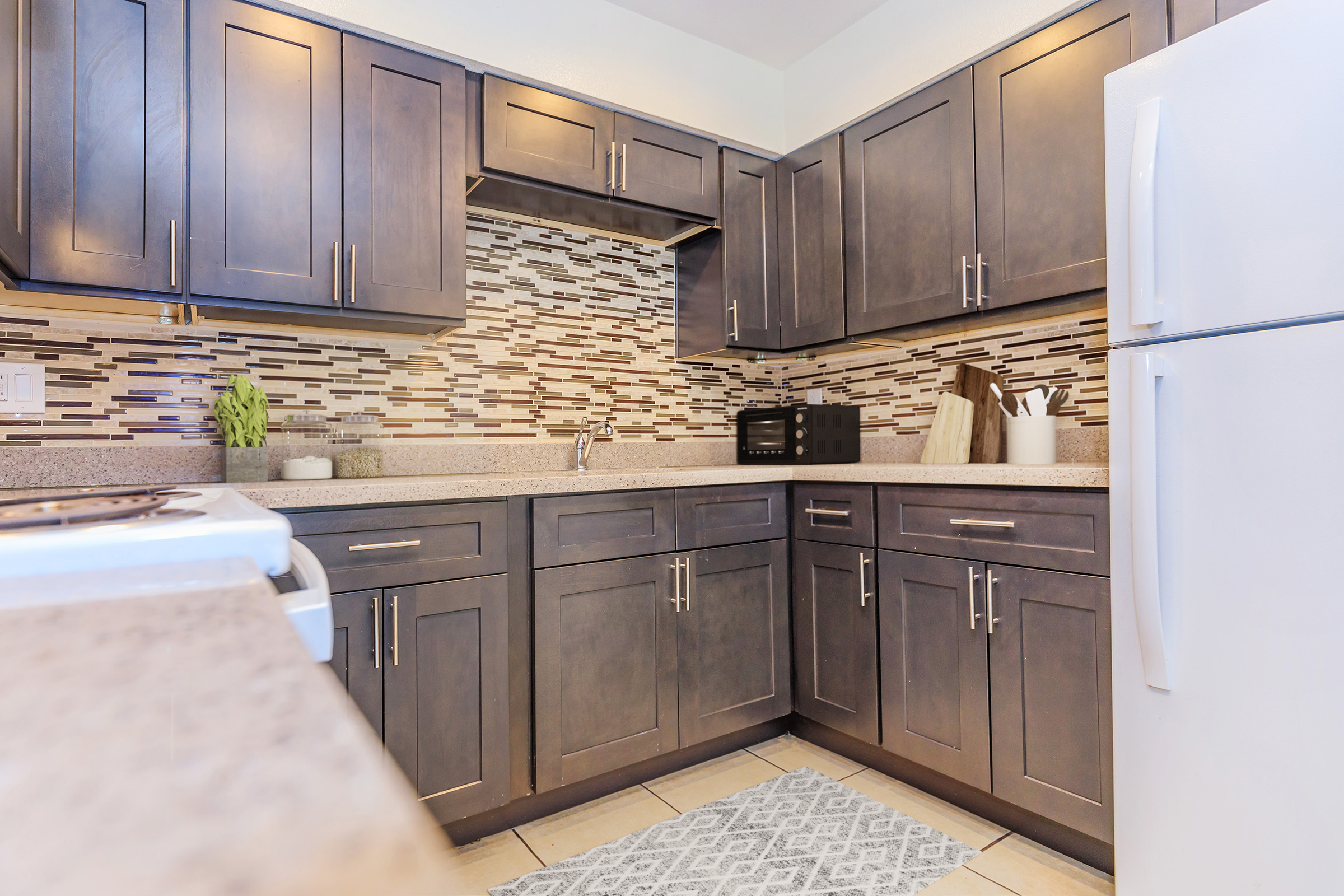 Modern kitchen featuring dark wood cabinets, a gray countertop, and a patterned backsplash. The space includes a microwave and utensils, with a light-colored rug on the floor. A white refrigerator is visible, creating a clean and contemporary look.