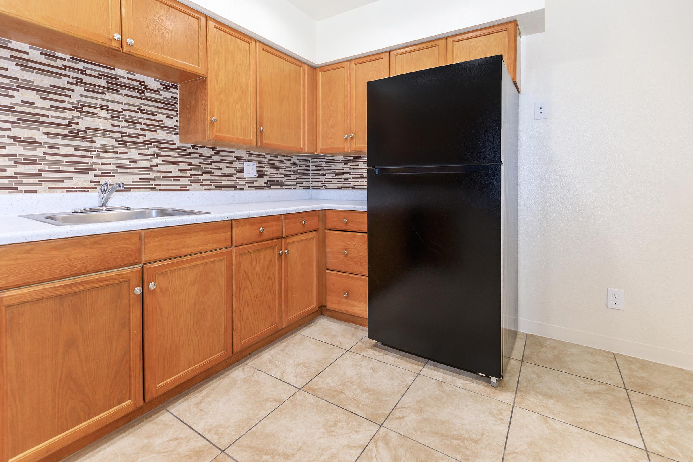 A modern kitchen featuring wooden cabinetry, a black refrigerator, and a tiled floor. The backsplash has a mosaic design with various shades and patterns. A stainless steel sink is installed in the countertop, creating a clean and functional cooking space.