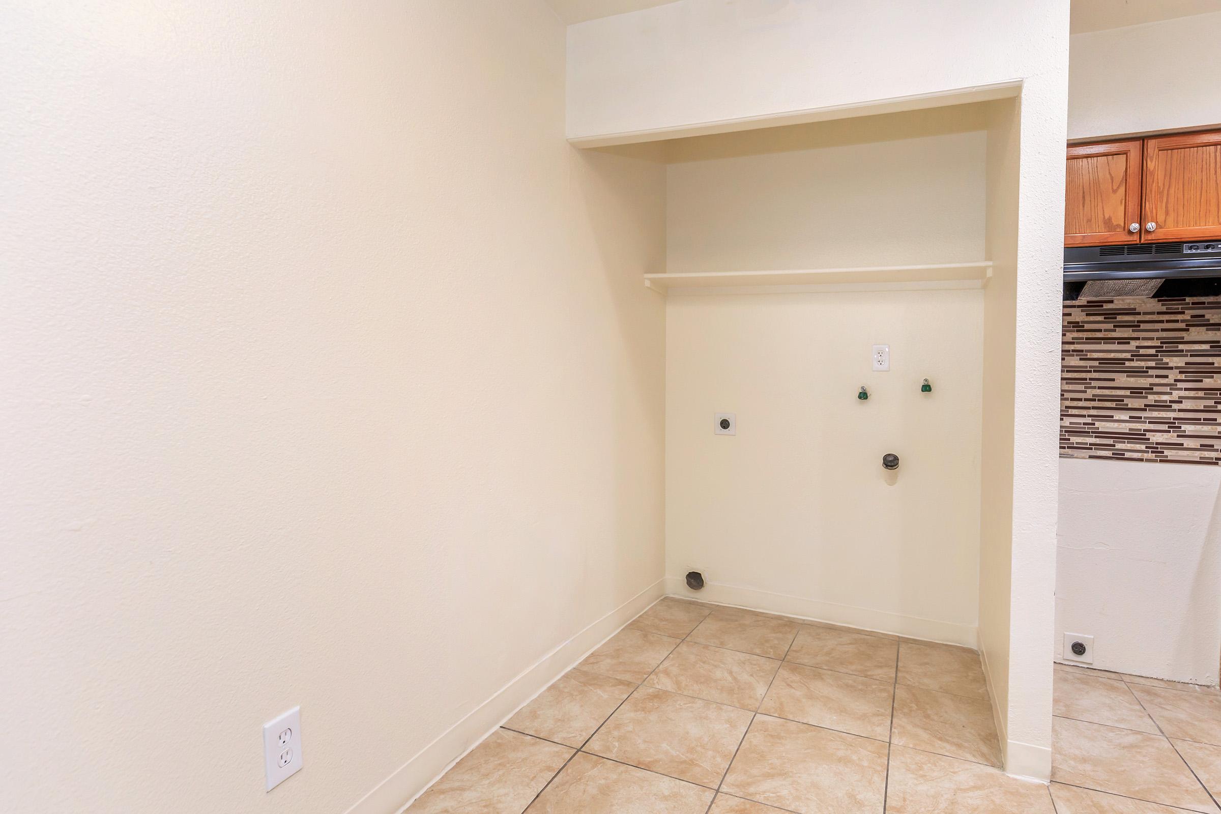 Empty laundry area with a tiled floor and light-colored walls. There is a shelf above a wall-mounted electrical outlet and plumbing fixtures. The area appears freshly painted and is adjacent to a kitchen with wooden cabinets.