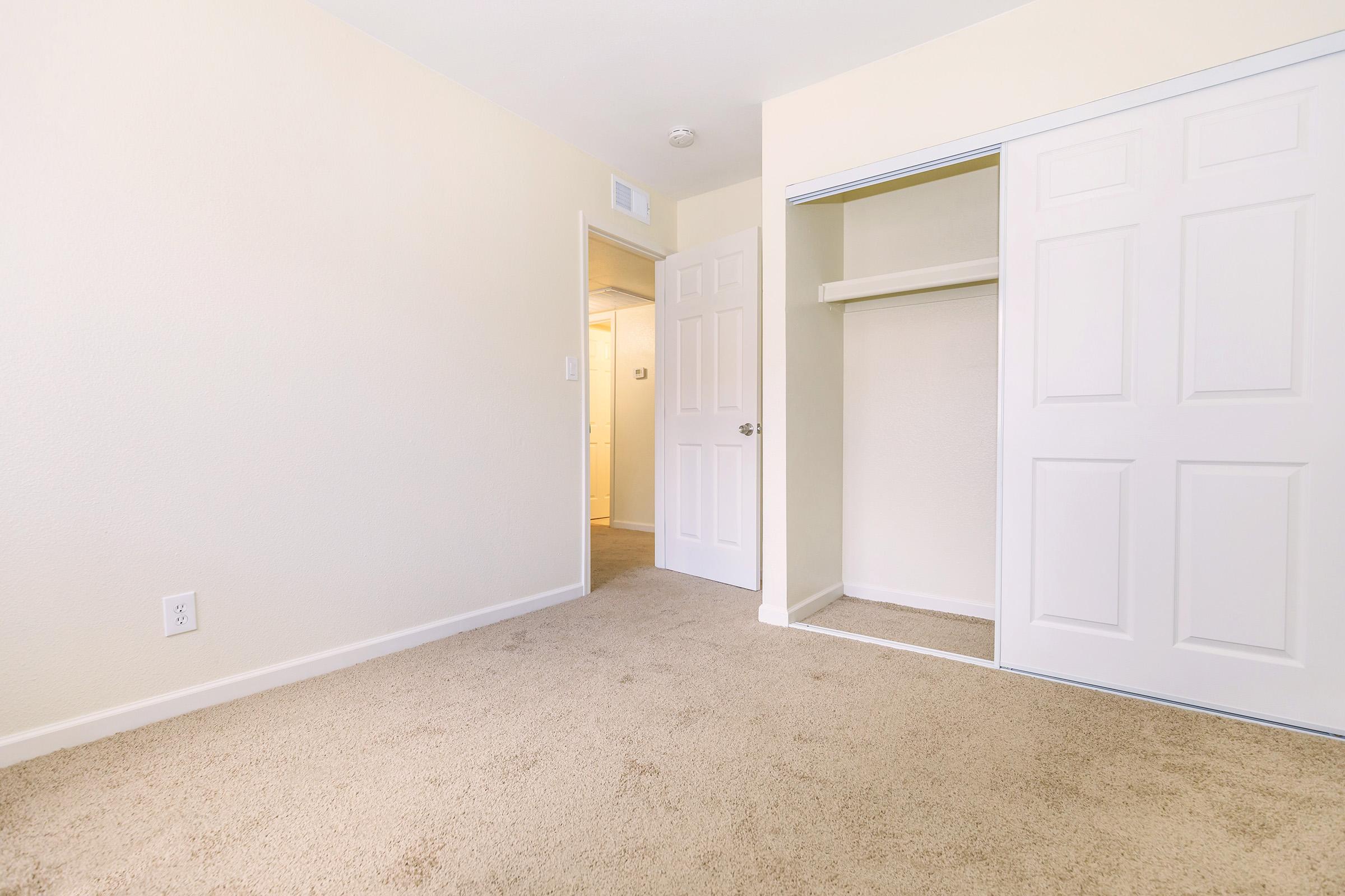 A spacious room with beige walls and carpet, featuring a closet with sliding doors. A doorway leads to another area, possibly a bathroom or hallway. The overall atmosphere is bright and inviting, with natural light enhancing the neutral tones.