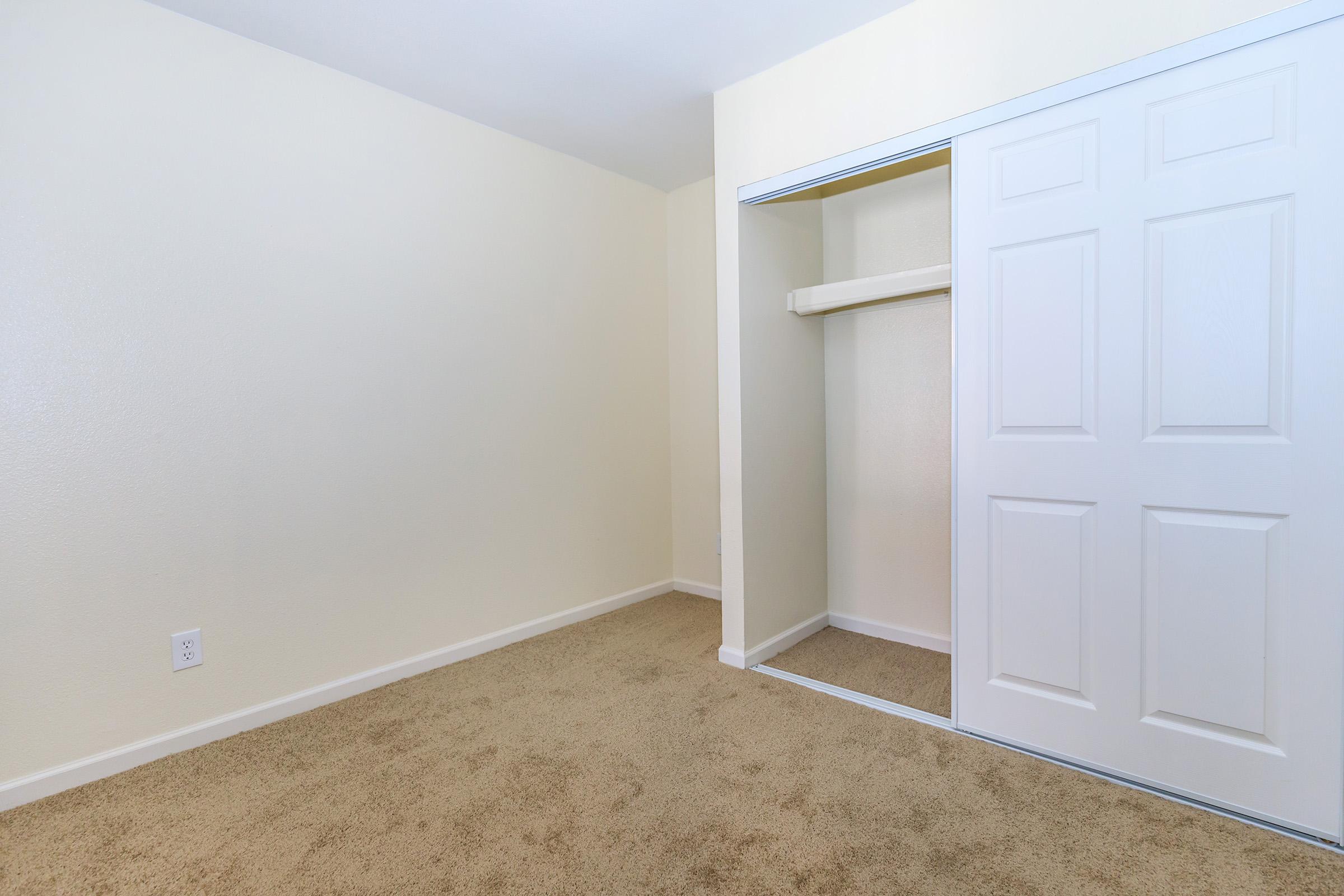 Empty room with beige walls and carpeted floor. There is a white closet with sliding doors on the right side of the image. The space is well-lit and minimalistic, suggesting it is suitable for various uses such as a bedroom or office.