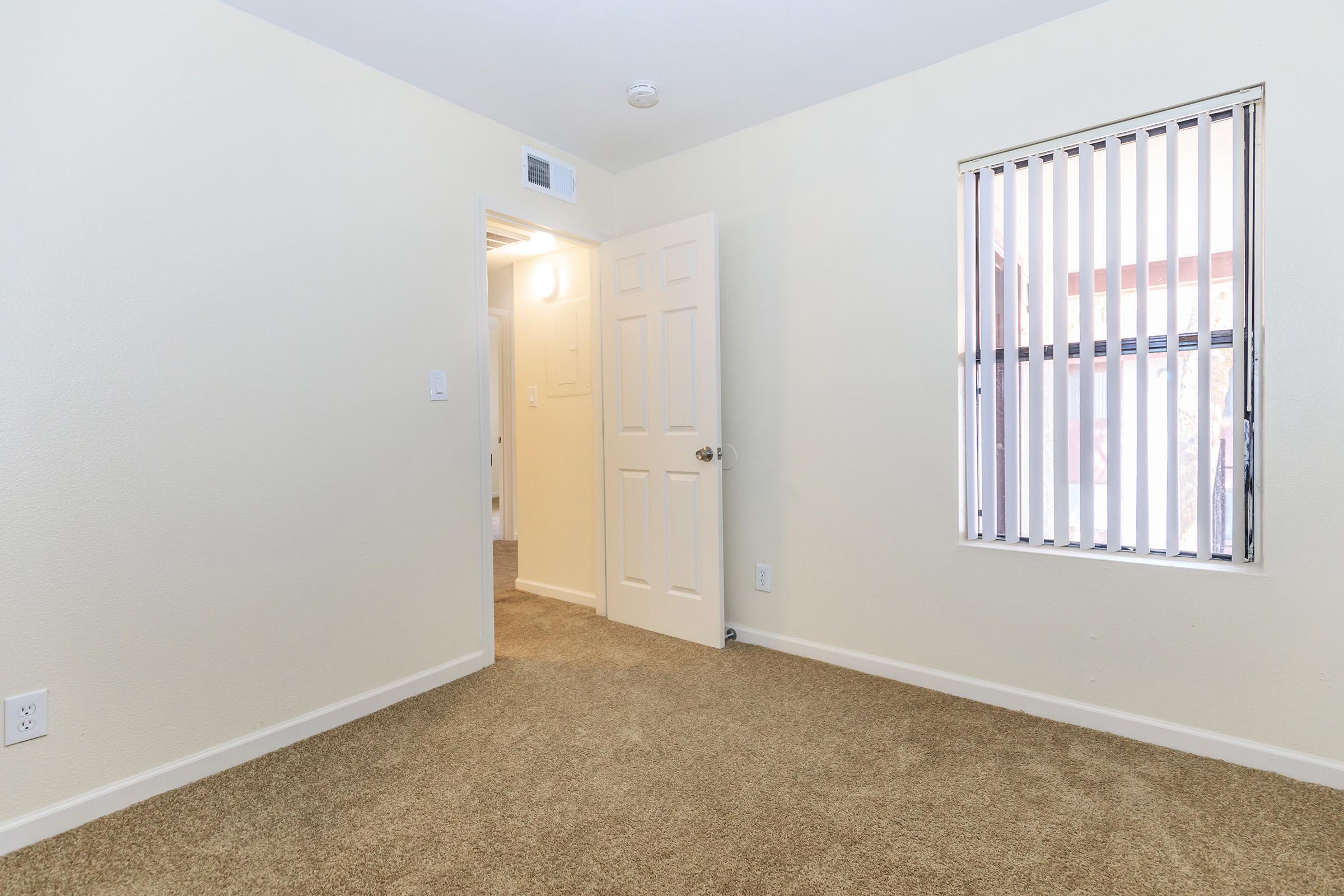 A light-colored room with beige carpeting and a beige wall. A closed door is visible, leading to a hallway, while a window with vertical blinds allows natural light. The overall atmosphere is spacious and airy, suitable for various purposes such as a bedroom or office.