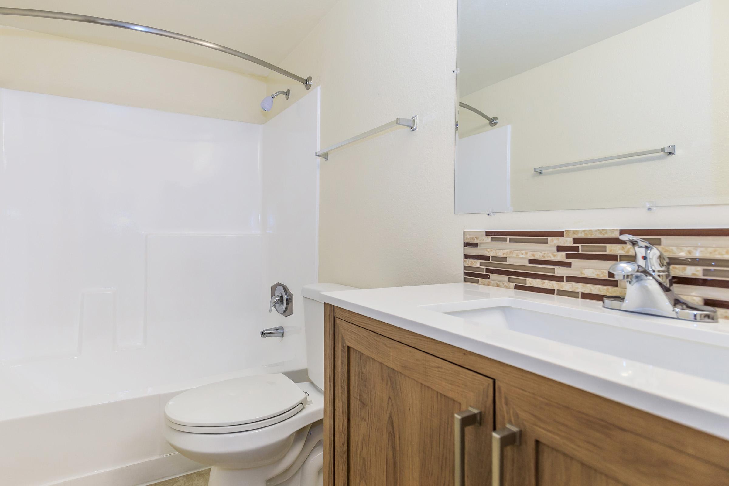 A clean, modern bathroom featuring a white bathtub with a shower, a wooden vanity with a sink, and a mirror. The walls are painted in a light shade, and the backsplash behind the sink has a decorative tile pattern.