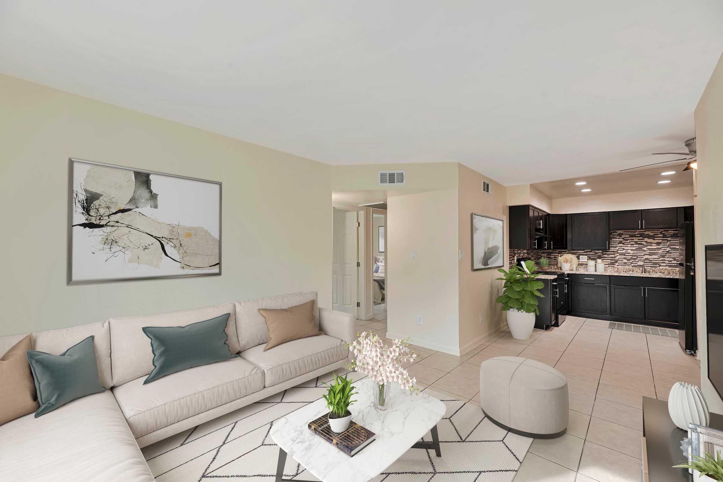 Modern living room featuring a light-colored sofa with decorative pillows, a coffee table with a floral arrangement, and a stylish area rug. The background showcases an open kitchen with dark cabinets and neutral walls, creating a warm and inviting atmosphere. Natural light enhances the space.