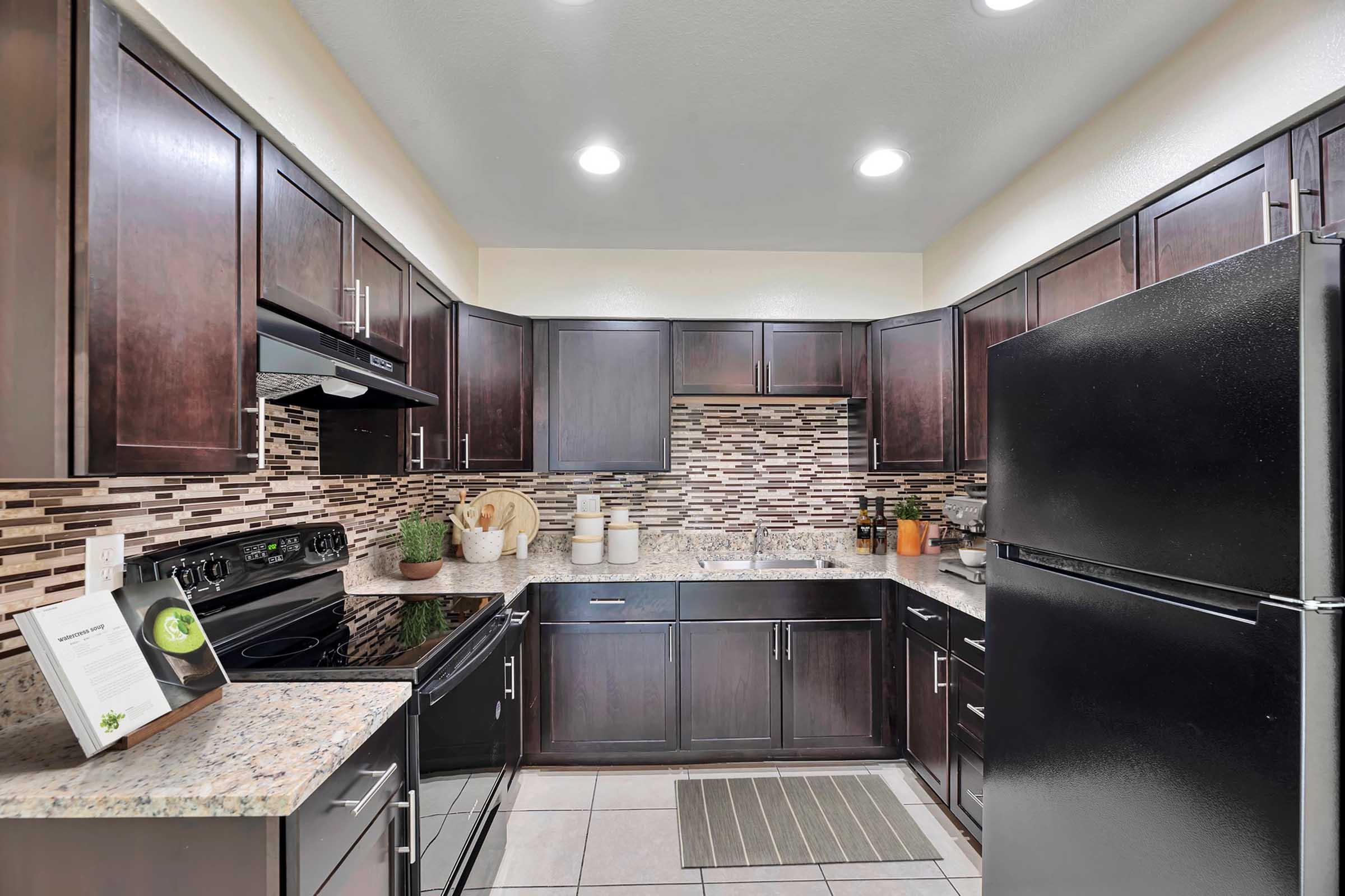 Modern kitchen with dark wood cabinets, a black refrigerator, and stainless steel appliances. The countertops are made of light-colored granite, and the backsplash features mixed brown tiles. A dish rack and potted plants are visible, along with a recipe book on the countertop. Well-lit with overhead lights.