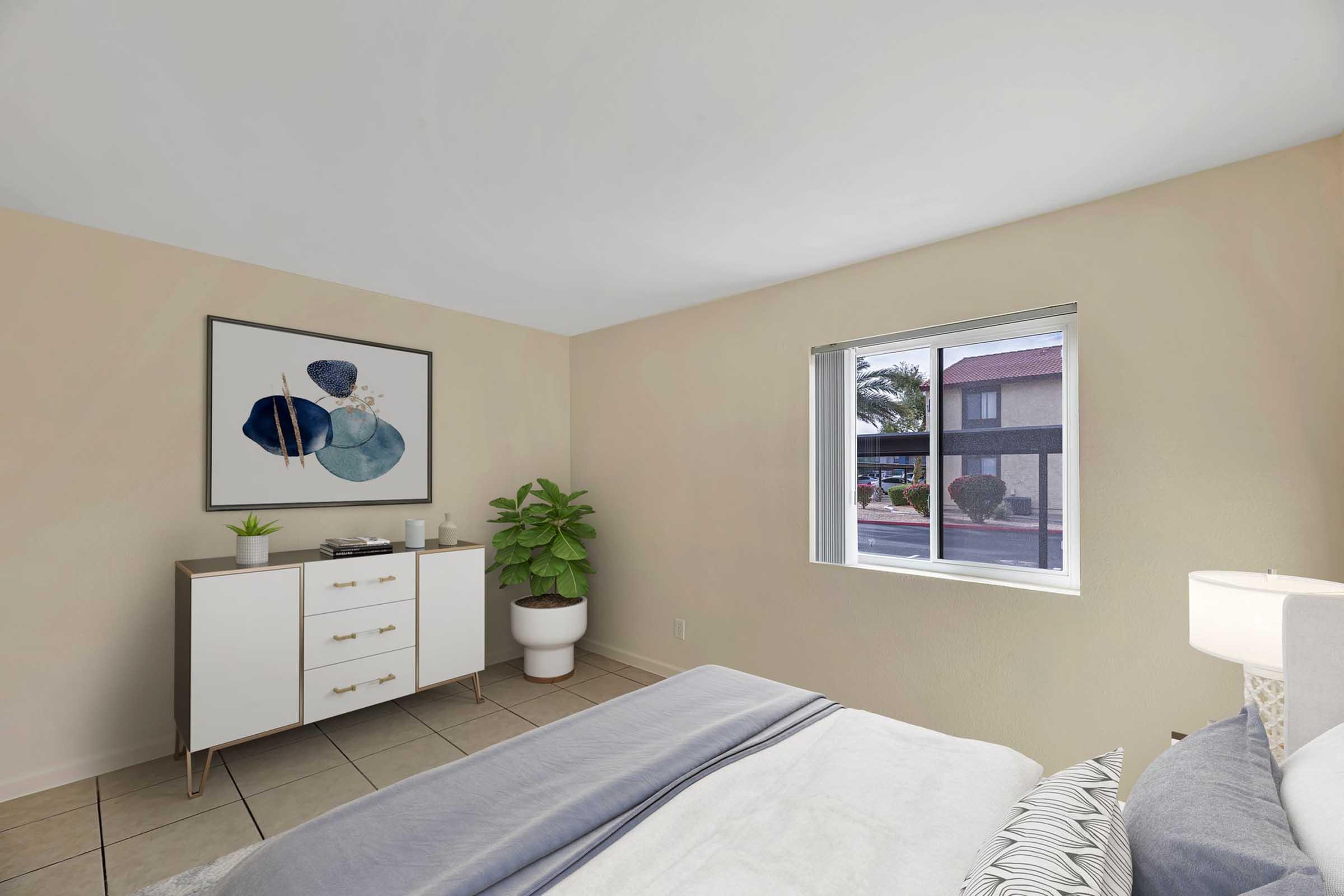 A cozy bedroom featuring light beige walls, a large window with outside views, a modern white dresser with gold handles, a green potted plant, and a decorative wall art piece with abstract shapes. The bed has a gray blanket and a bedside lamp, creating a warm and inviting atmosphere.