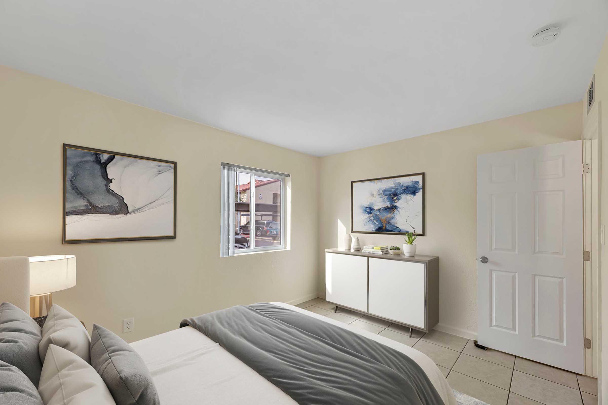 A cozy bedroom featuring a bed with gray linens, a bedside lamp, and two abstract paintings on the wall. There's a window letting in natural light, a small green plant on a modern white dresser, and a closed door leading to another space. The room has light-colored walls and tile flooring.